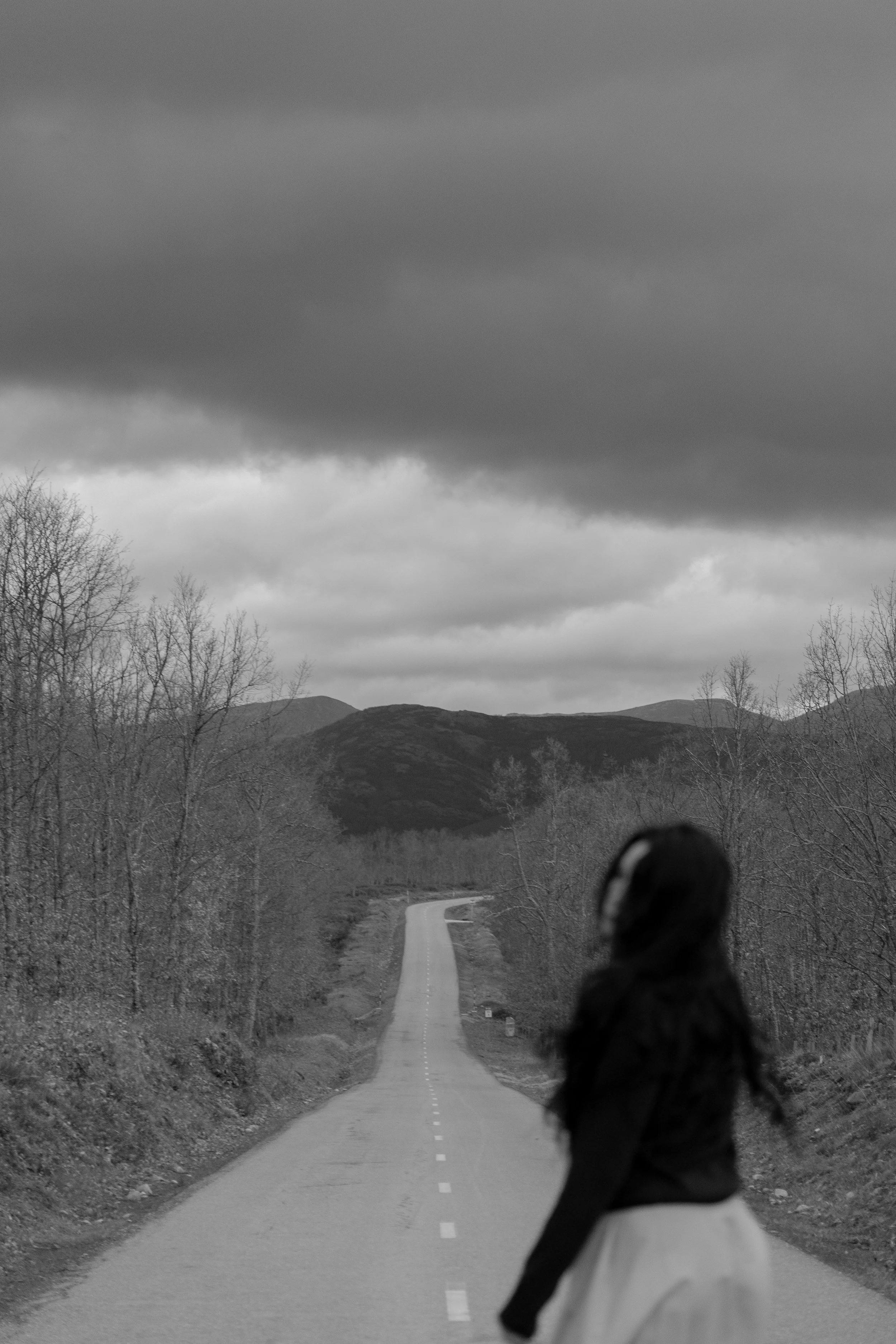 A person standing on a deserted road through a rural landscape with mountains and leafless trees, under dark, cloudy skies.
