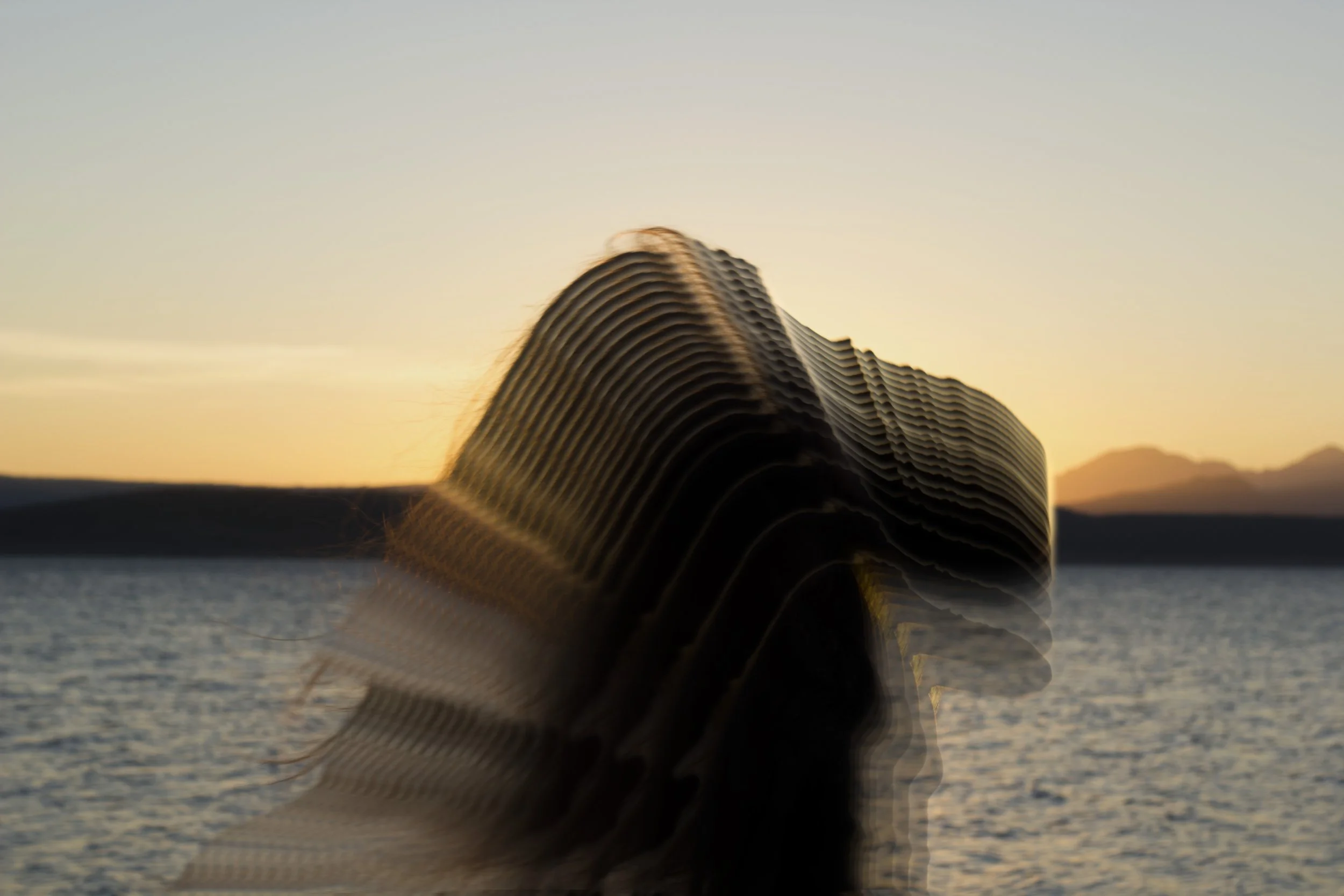 Blurry silhouette of a person with long hair standing near a lake or sea at sunset or sunrise.
