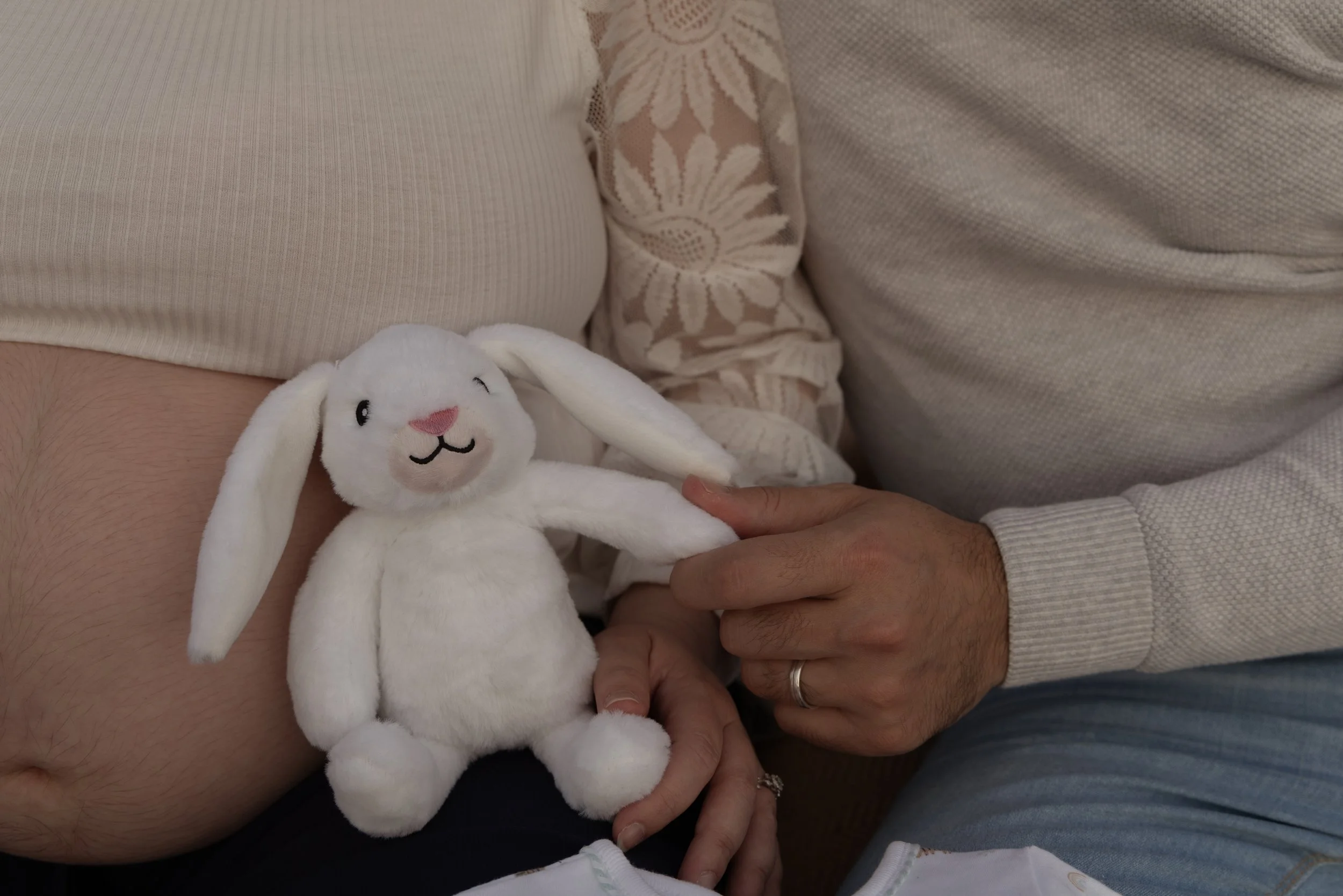 A stuffed white bunny with floppy ears, pink nose, and embroidered face is held by a person with a ring on their finger, sitting close to another person wearing a beige embroidered top and light-colored sweater in Cramong beach