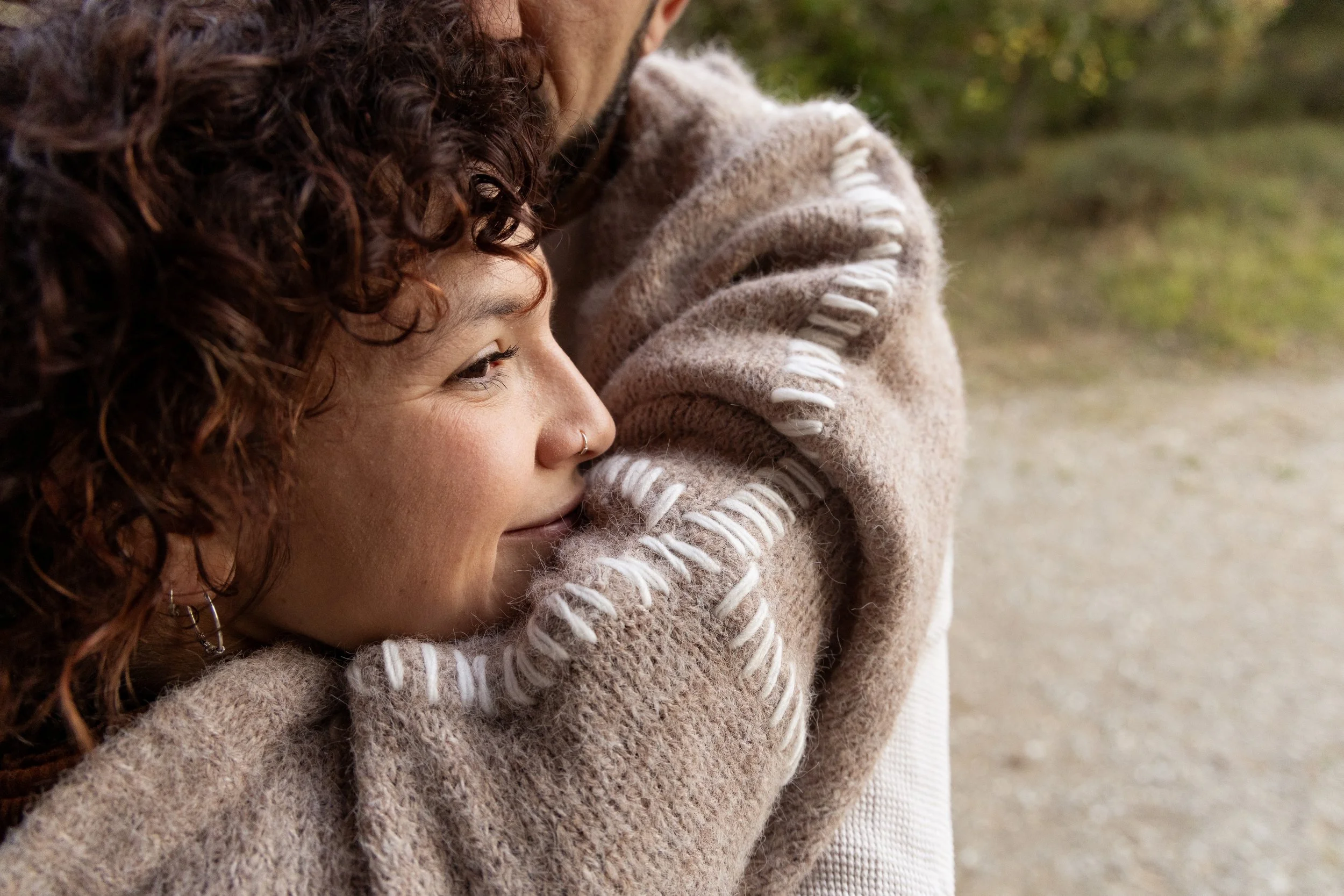 Close-up of a smiling woman with curly hair and a nose ring, wrapped in a cozy beige sweater, outdoors with blurred greenery in the background.
