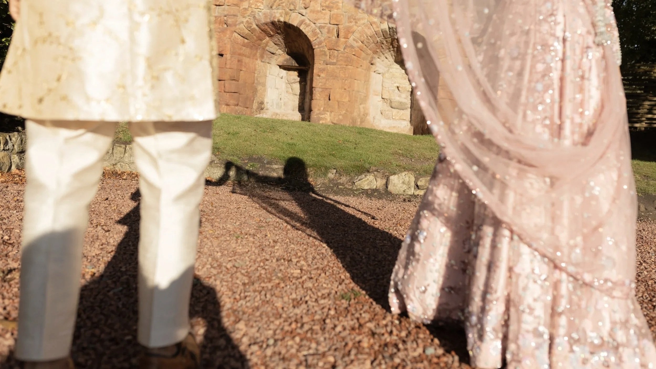 Close-up of two people holding hands, standing on gravel path with an ancient brick archway in the background, sunlight casting shadows.