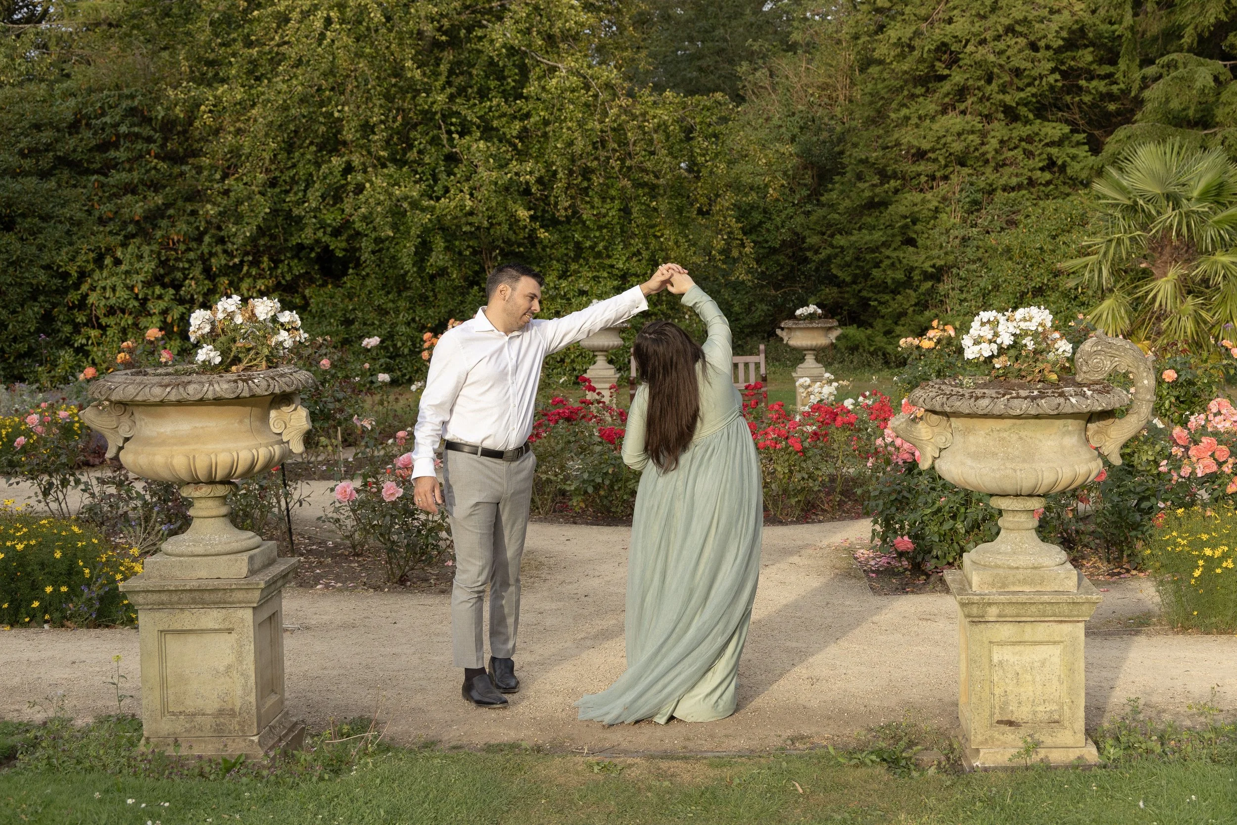 A man and woman dancing in a garden surrounded by colorful flowers and greenery, with stone planters and a bench in the background.