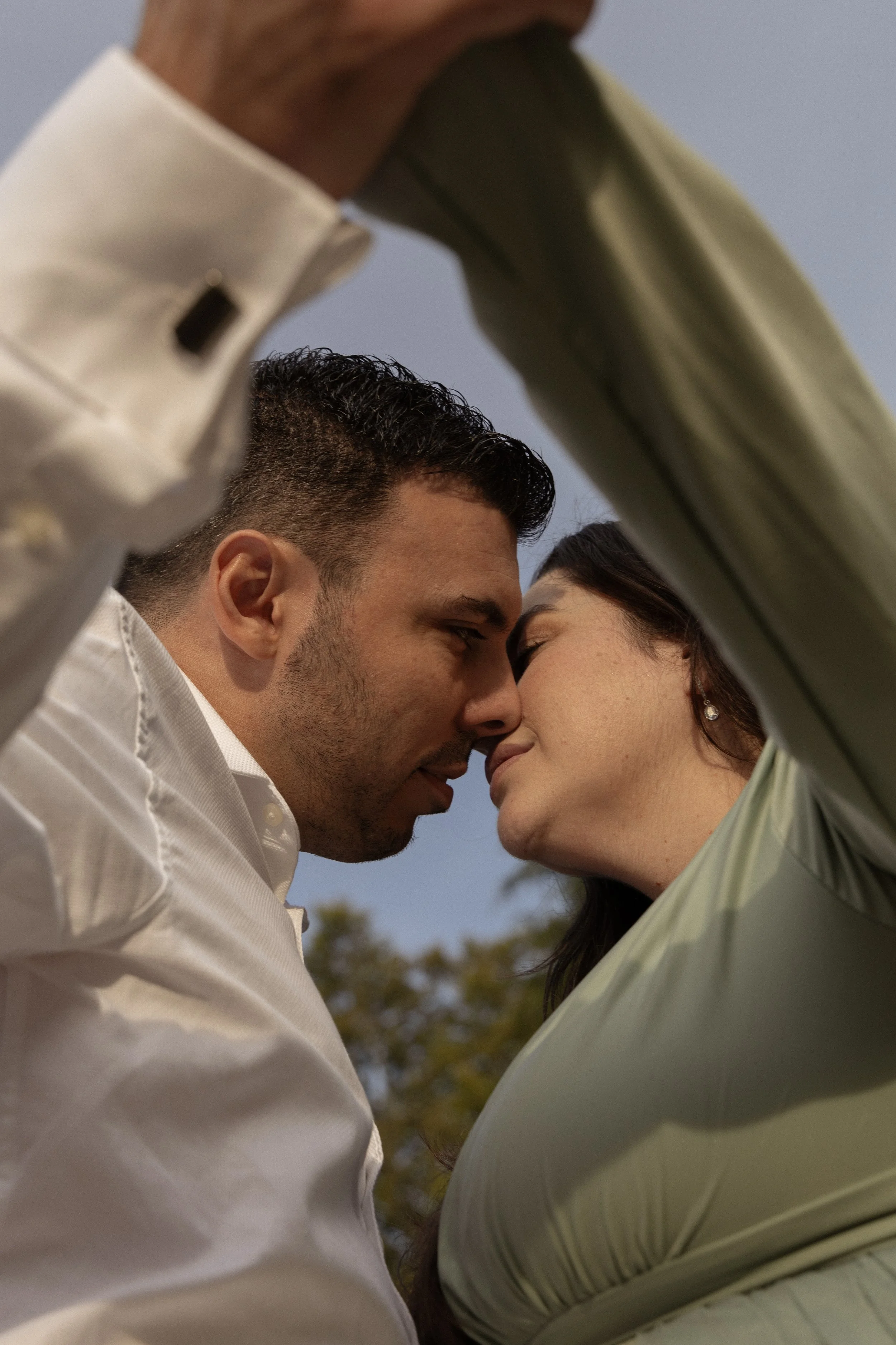 A close-up of a couple touching foreheads with their eyes closed, sharing an intimate moment outdoors with a blurry background of trees and a blue sky.