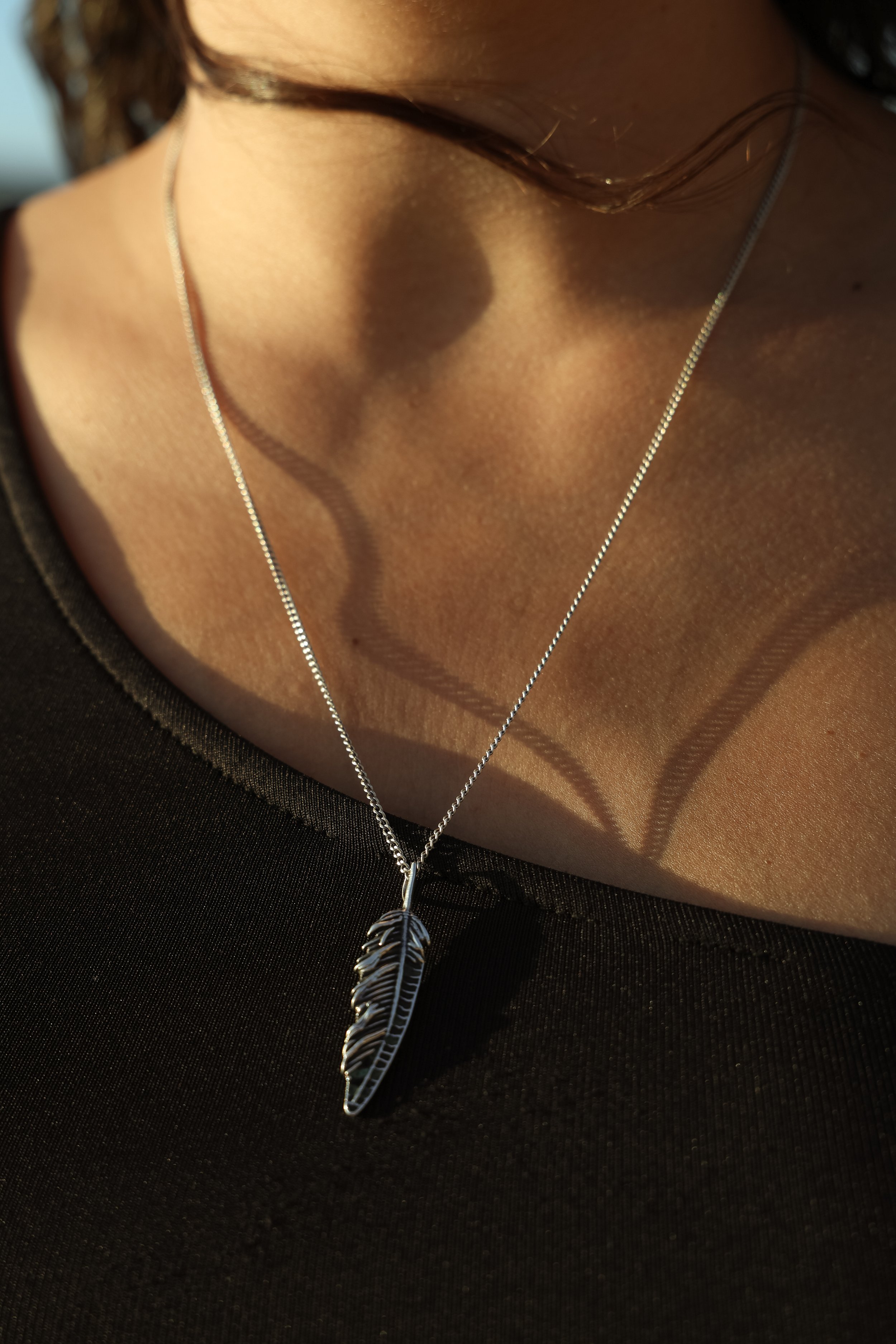 Close-up of a person wearing a silver necklace with a feather pendant, casting shadows on their skin and black shirt.