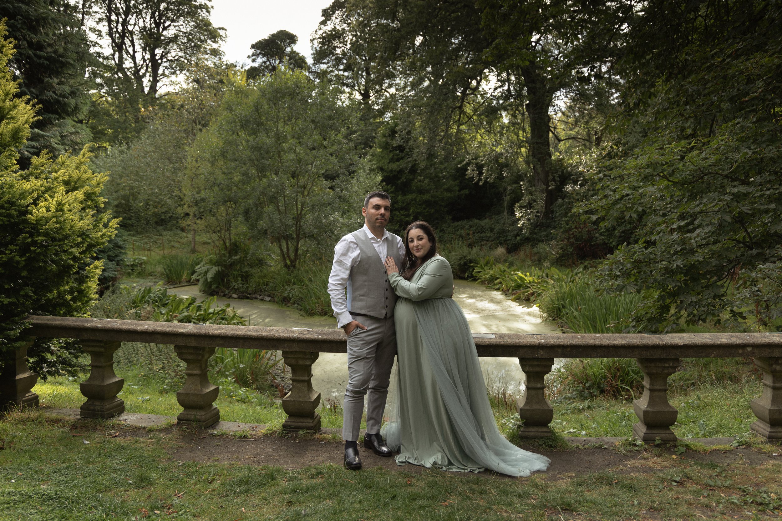 A couple in formal attire standing by a stone railing in a lush garden surrounded by trees and greenery in Lauriston Castle in Edinburgh