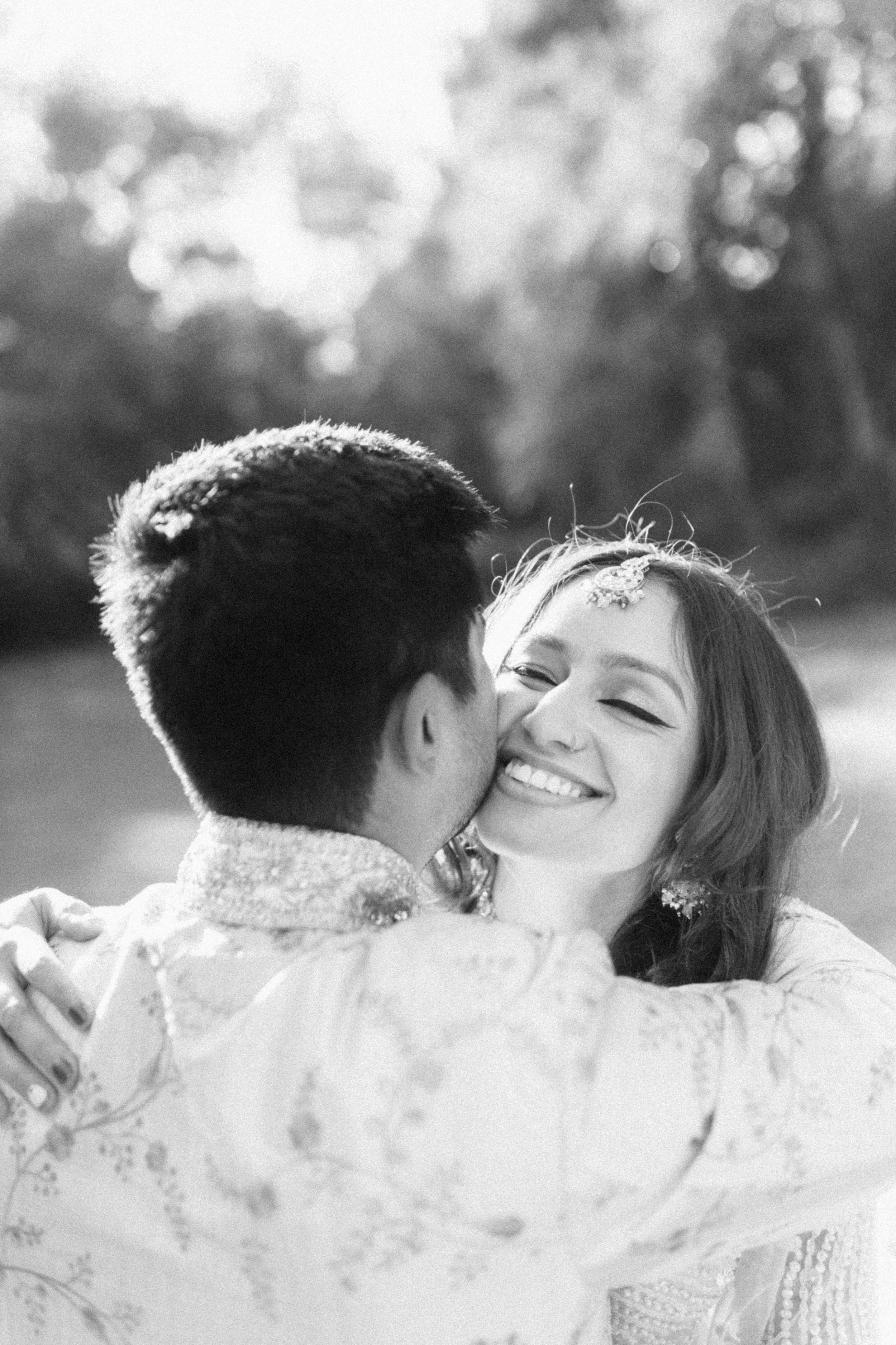 A couple hugging outdoors, with the woman smiling and the man kissing her cheek, in a black and white photograph.