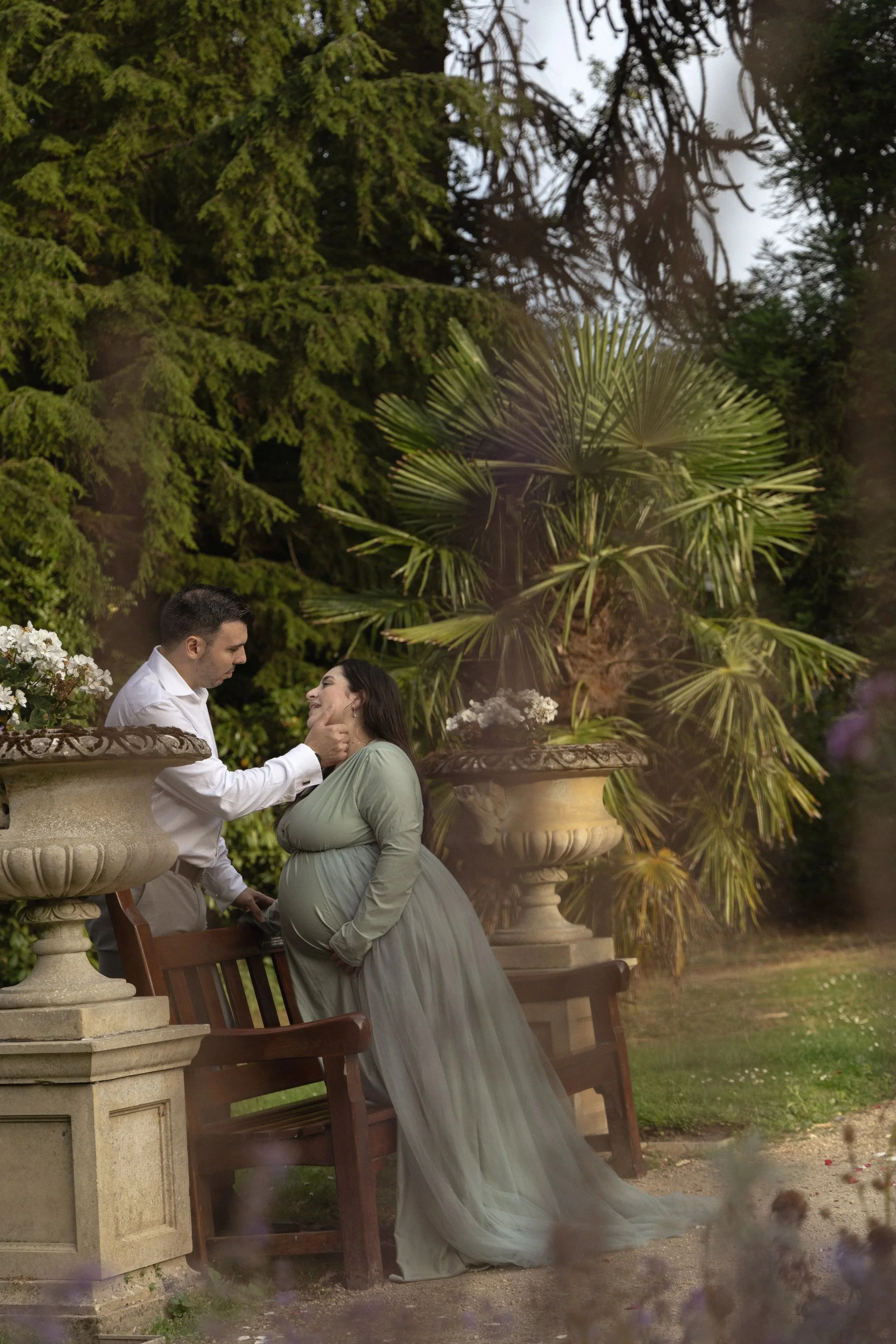 A man and pregnant woman sharing a tender moment in a lush garden, with large potted plants and green trees in the background.