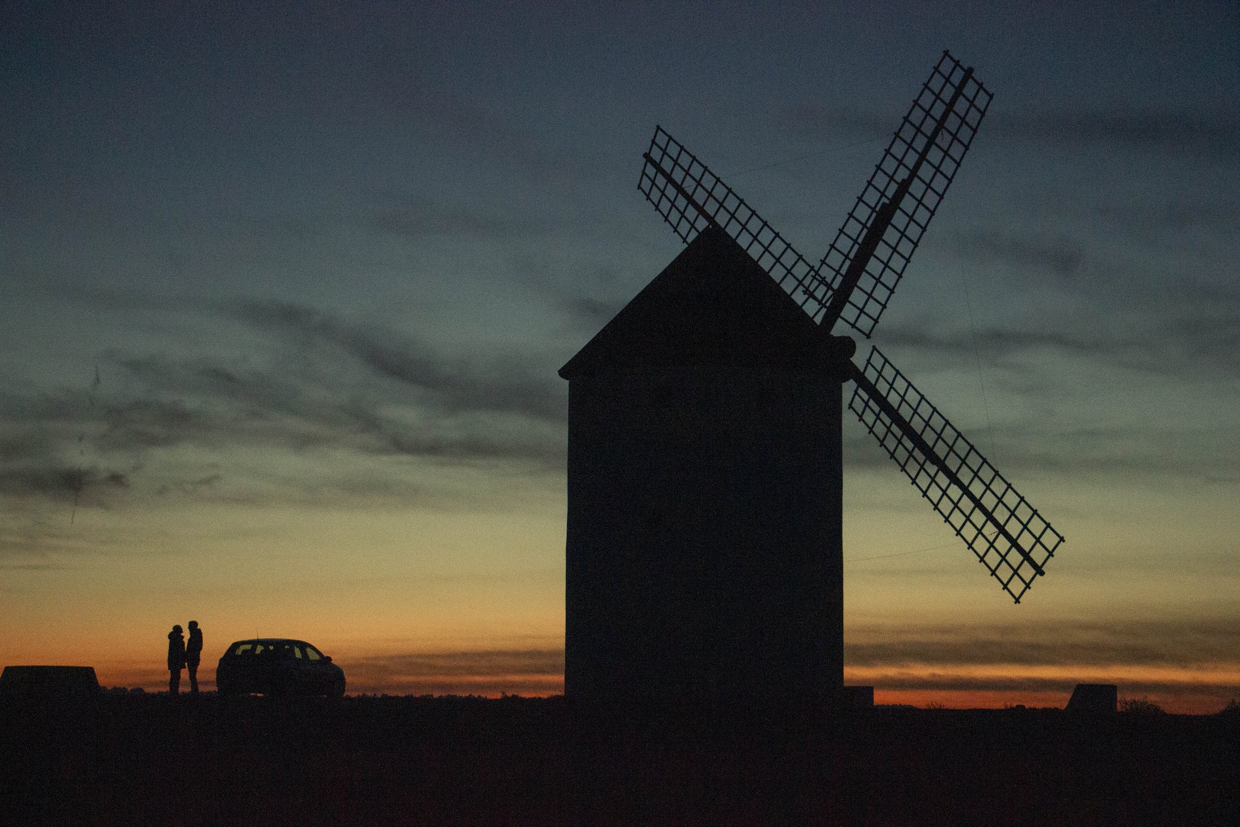 Silhouette of a windmill near sunset with a couple and car in the distance.