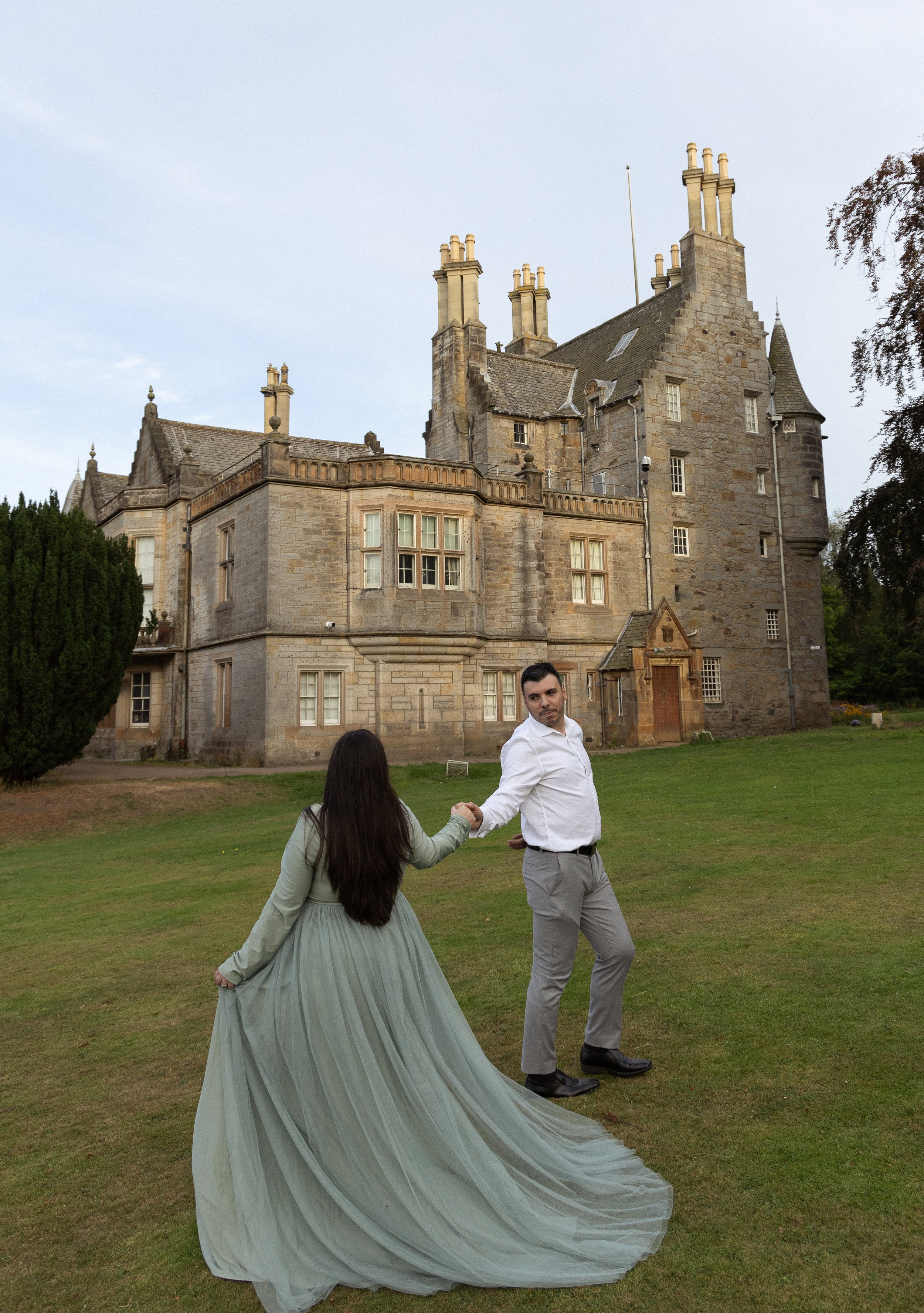 A couple dancing outdoors on a grassy area in front of thehistoric stone castle of Lauriston Caslte with multiple chimneys and turrets, under a blue sky.