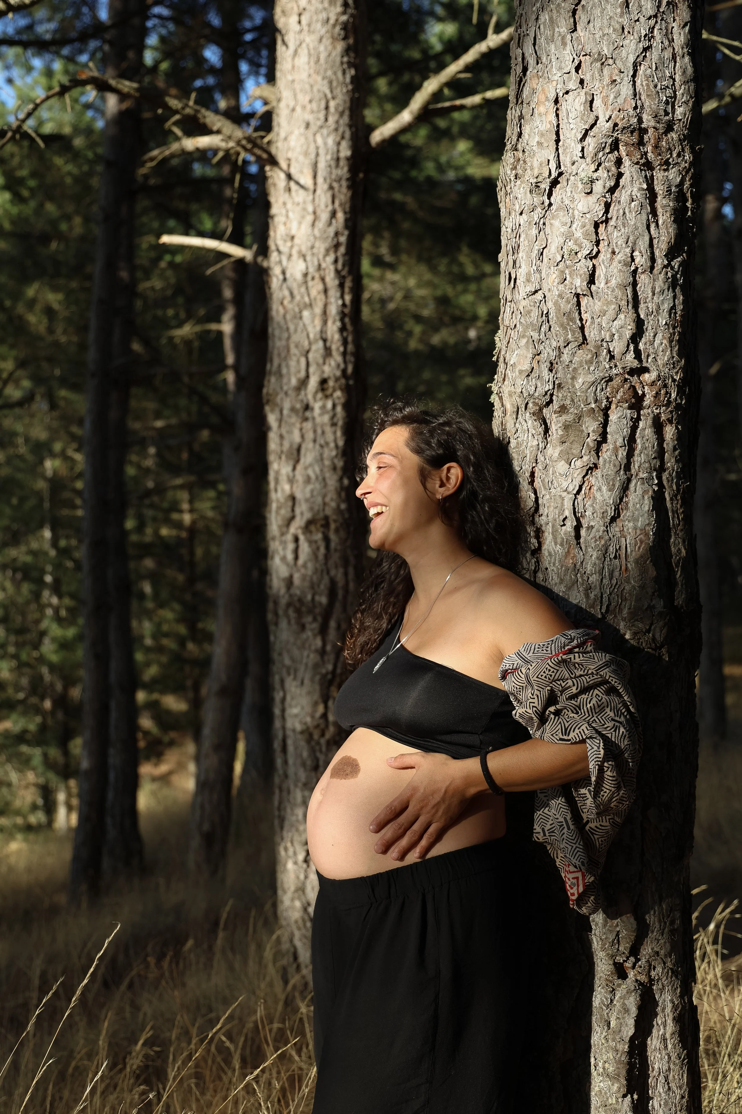 A pregnant woman with long dark curly hair leaning against a tree in a forest, smiling with her eyes closed and hand on her belly, sunlight shining on her face and belly.
