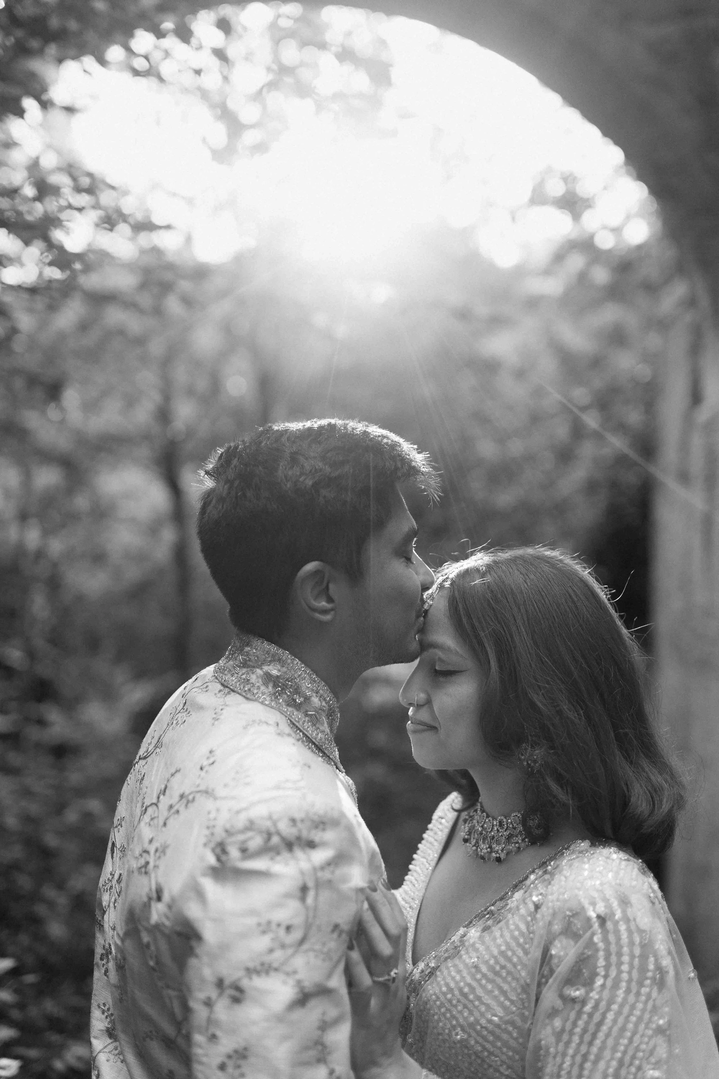 A couple in traditional Indian attire sharing a tender moment outdoors, with the man kissing the woman's forehead under sunlight filtering through trees.