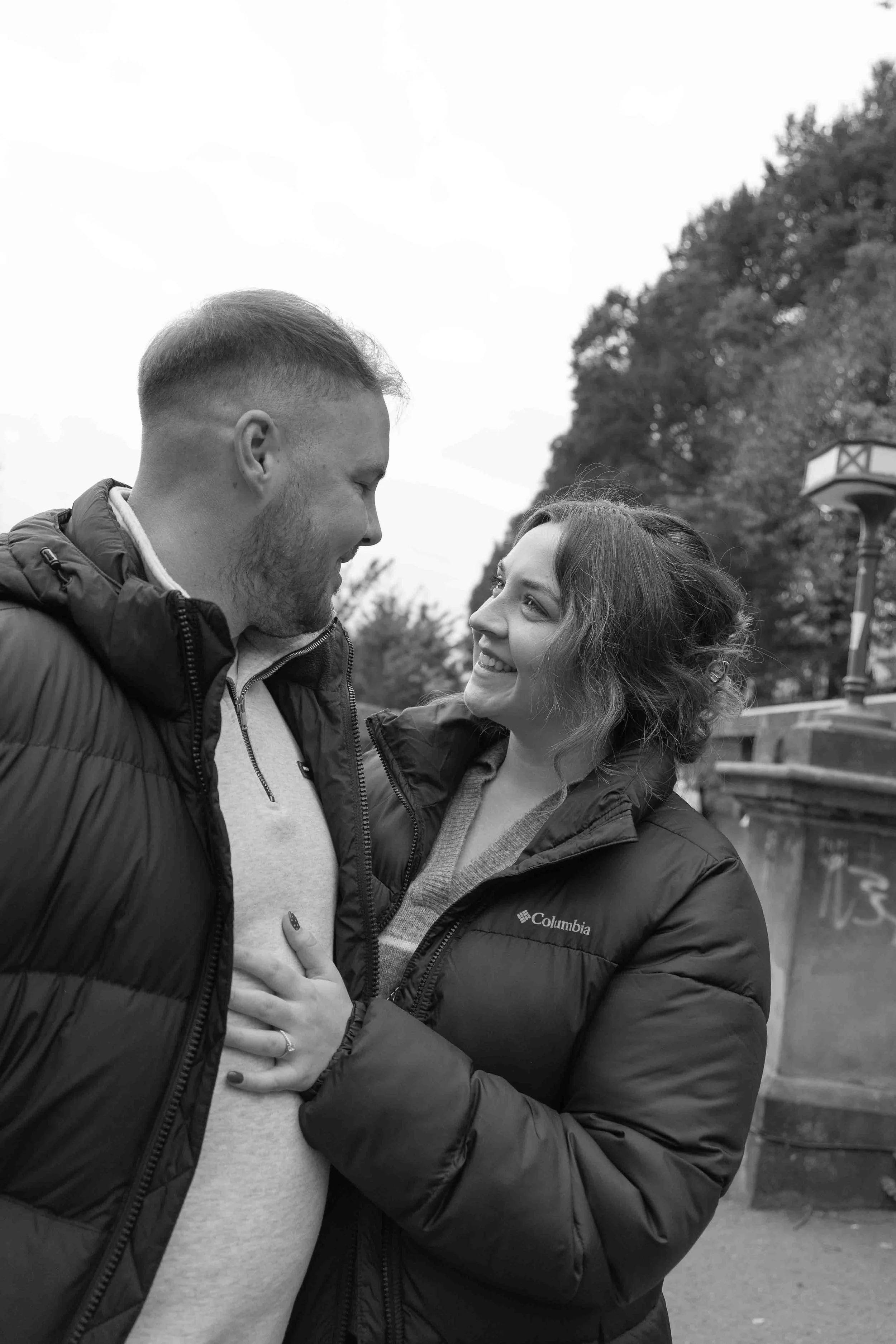 A smiling couple outdoors, looking at each other affectionately, dressed in warm jackets, with trees and a lamppost in the background.