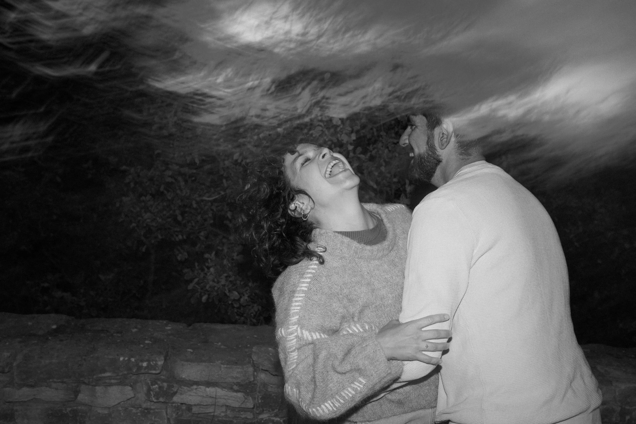 A woman and a man smiling and laughing, facing each other outdoors at night, with a cloudy sky overhead.