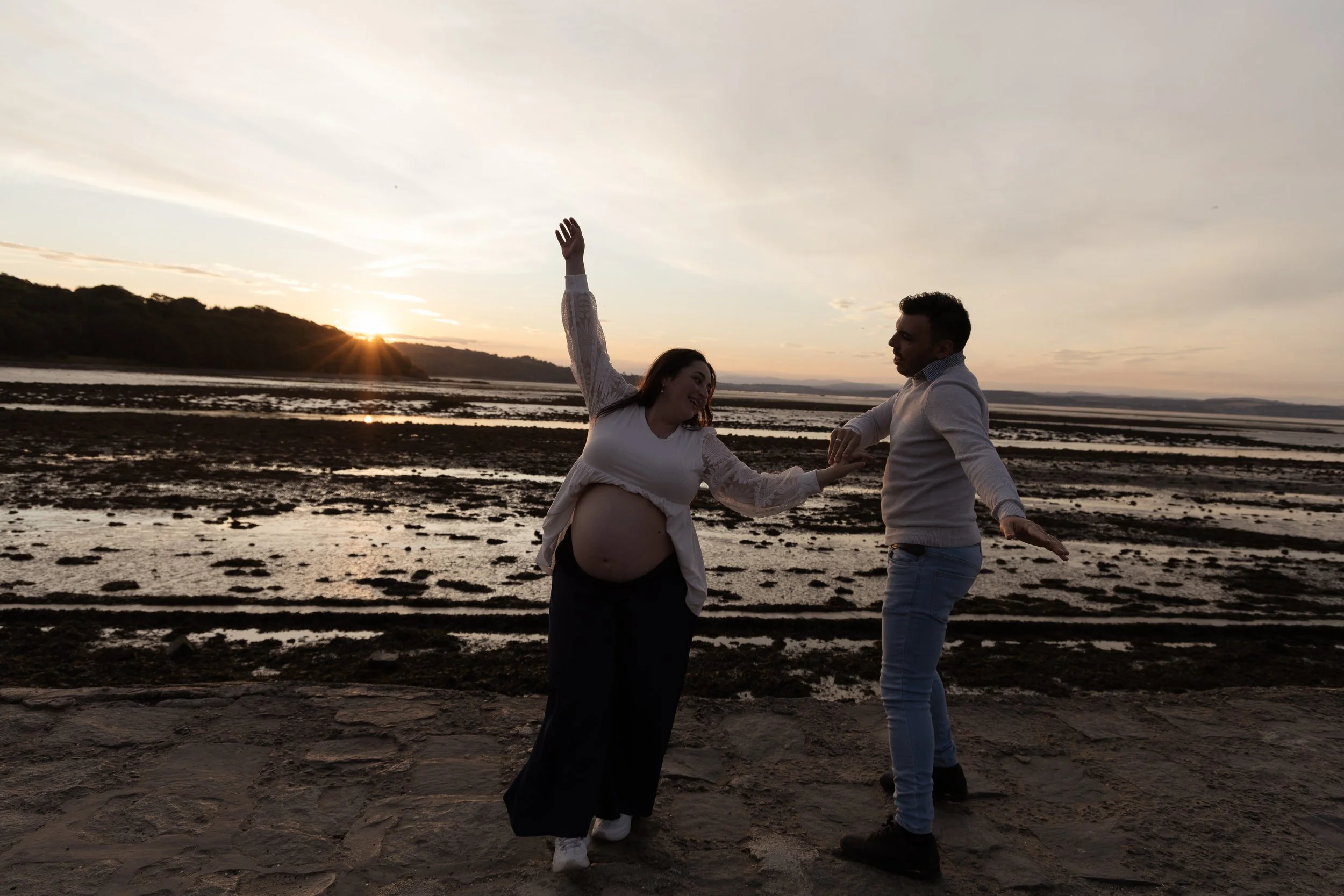 A pregnant woman and a man dancing on a beach during sunset, holding hands and smiling.