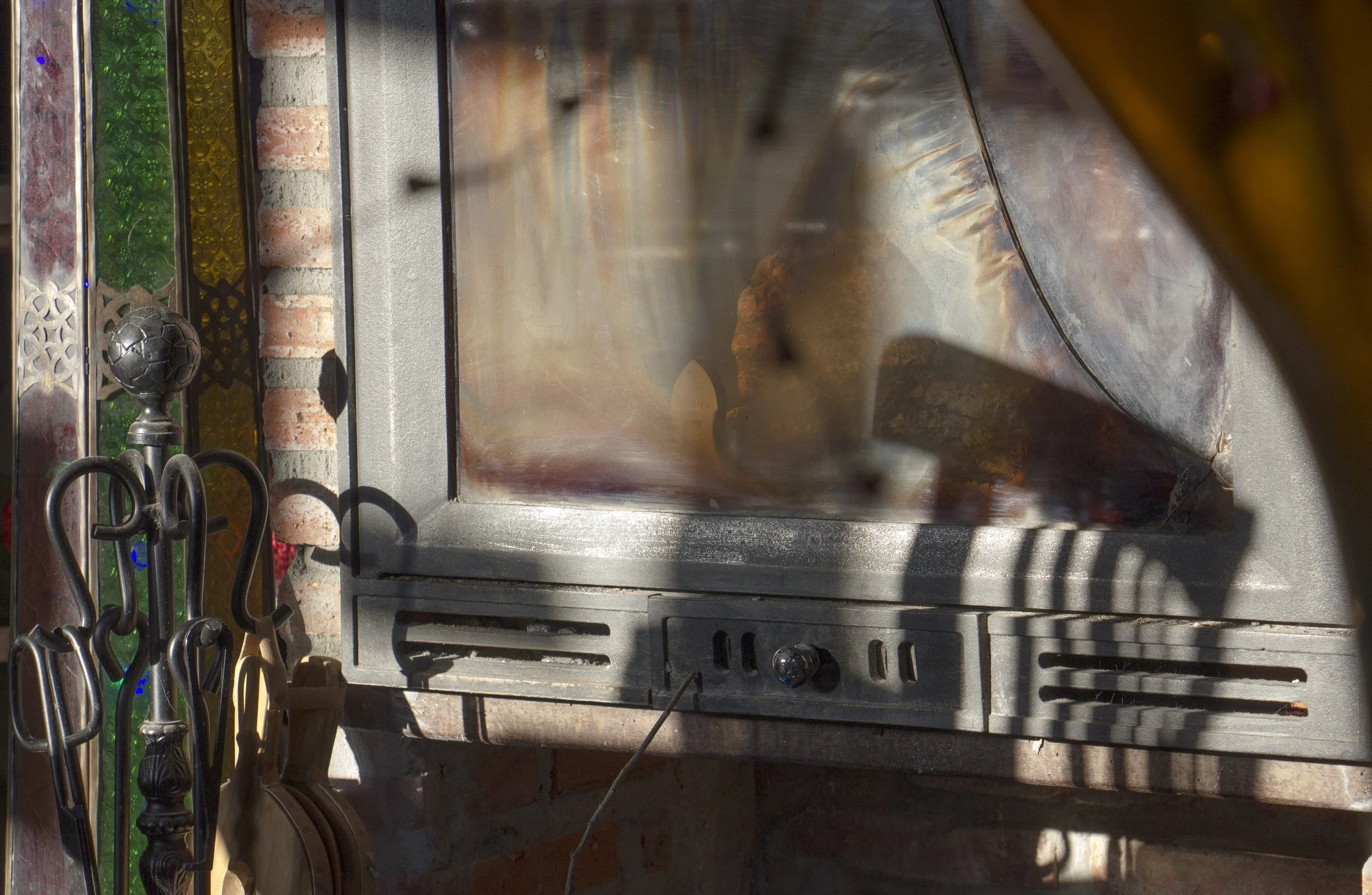 An outdoor fireplace or heating stove with glass door reflecting sunlight, surrounded by various metal tools and utensils hanging nearby, against a brick wall.