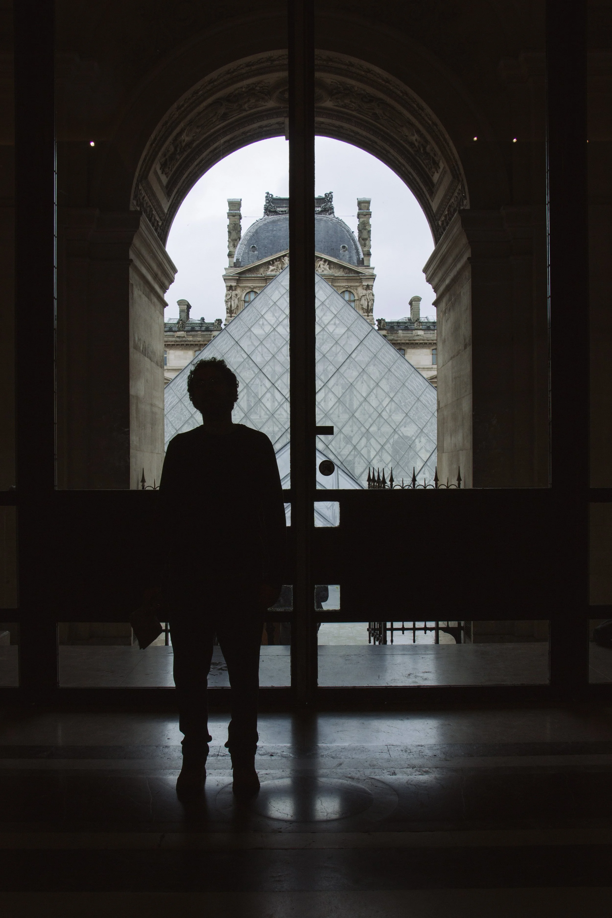 Silhouette of a person standing in front of a glass door with the Louvre Museum pyramid and classical building in the background.