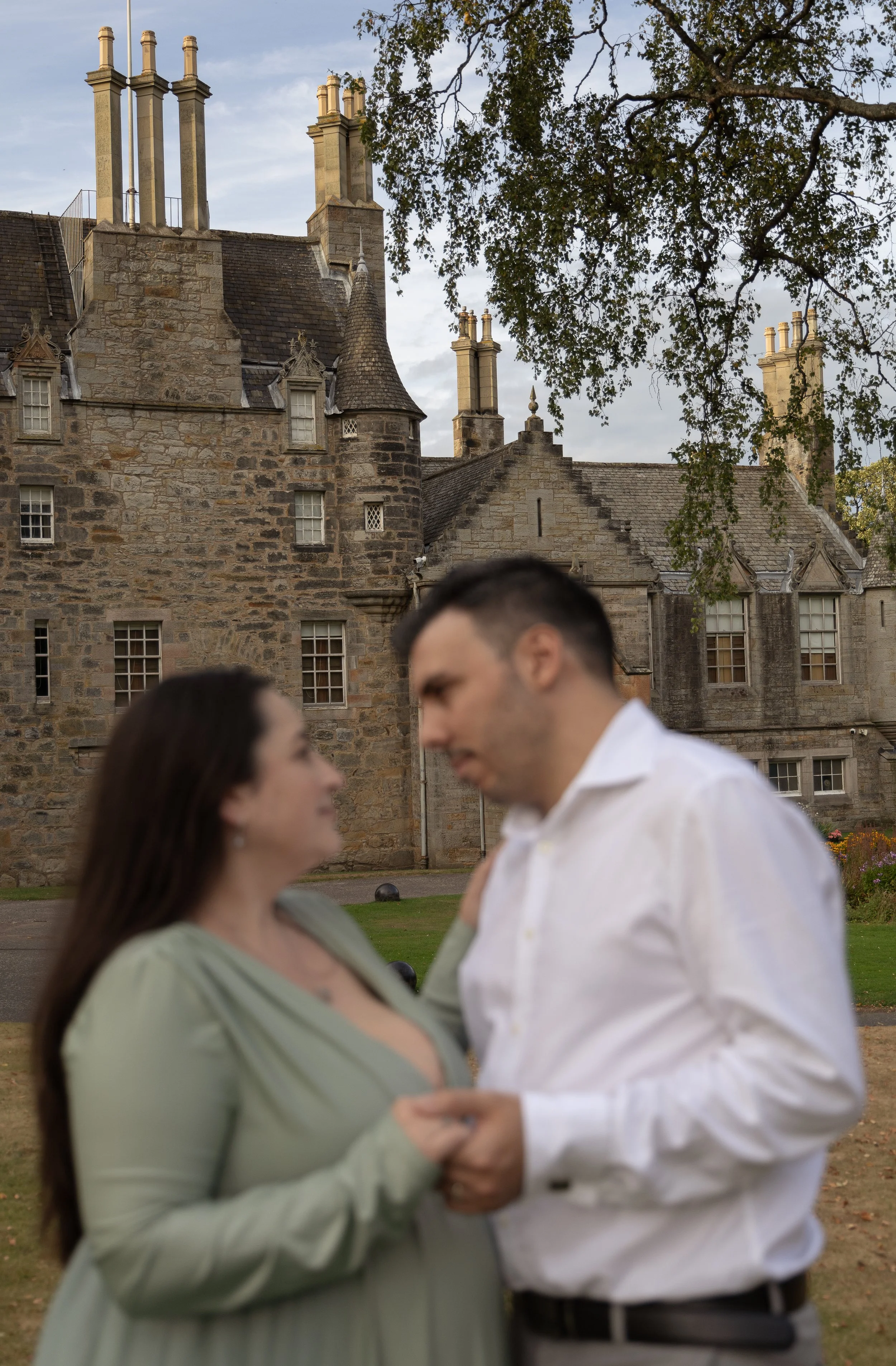 A blurry couple dancing together outdoors in front of a historic stone building with multiple chimneys and a tree with green leaves.