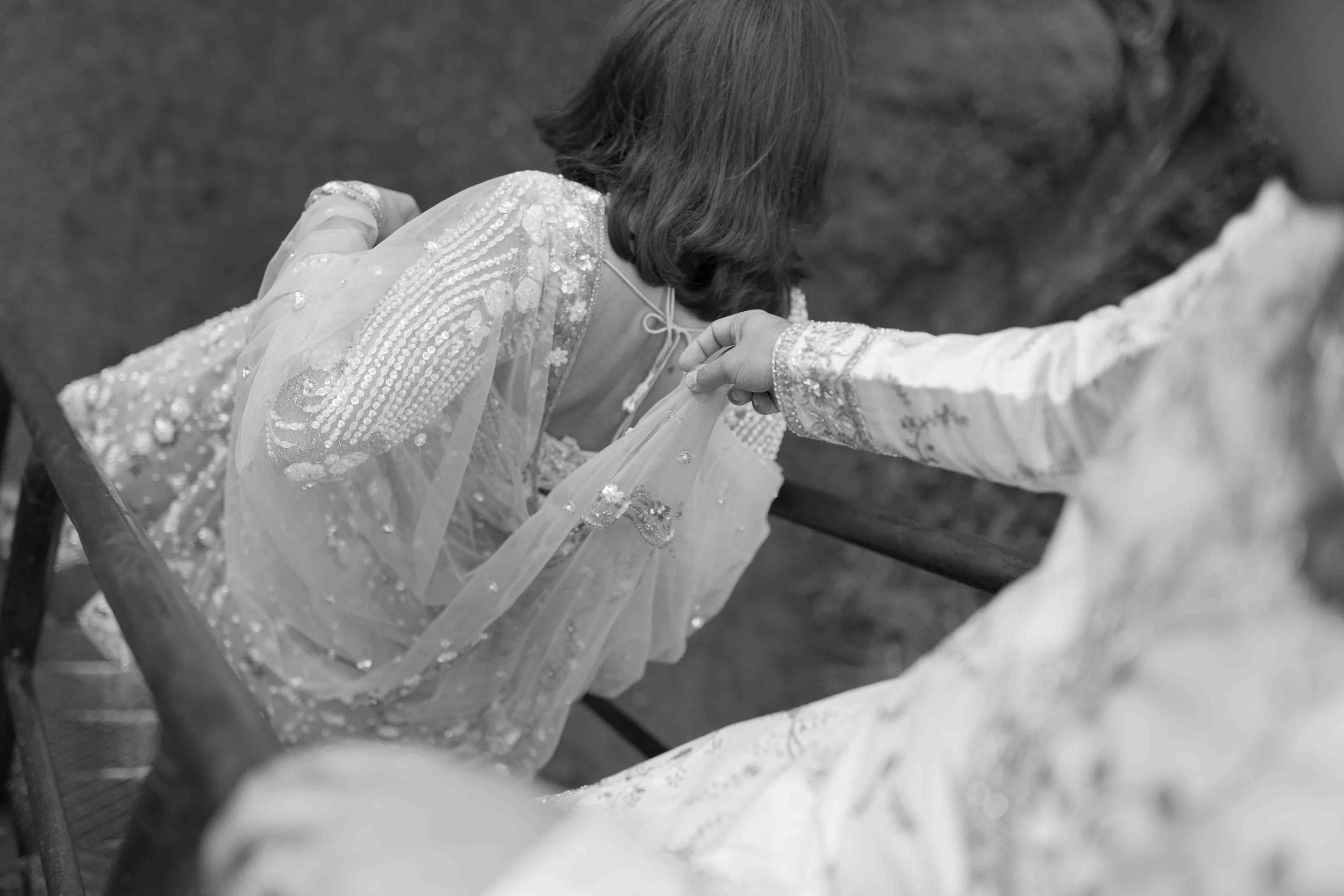 A woman in a heavily embroidered dress is sitting on a bench, leaning forward, with another person helping her adjust her dress or jewelry. The scene appears intimate and detailed, with focus on the traditional or ceremonial attire.