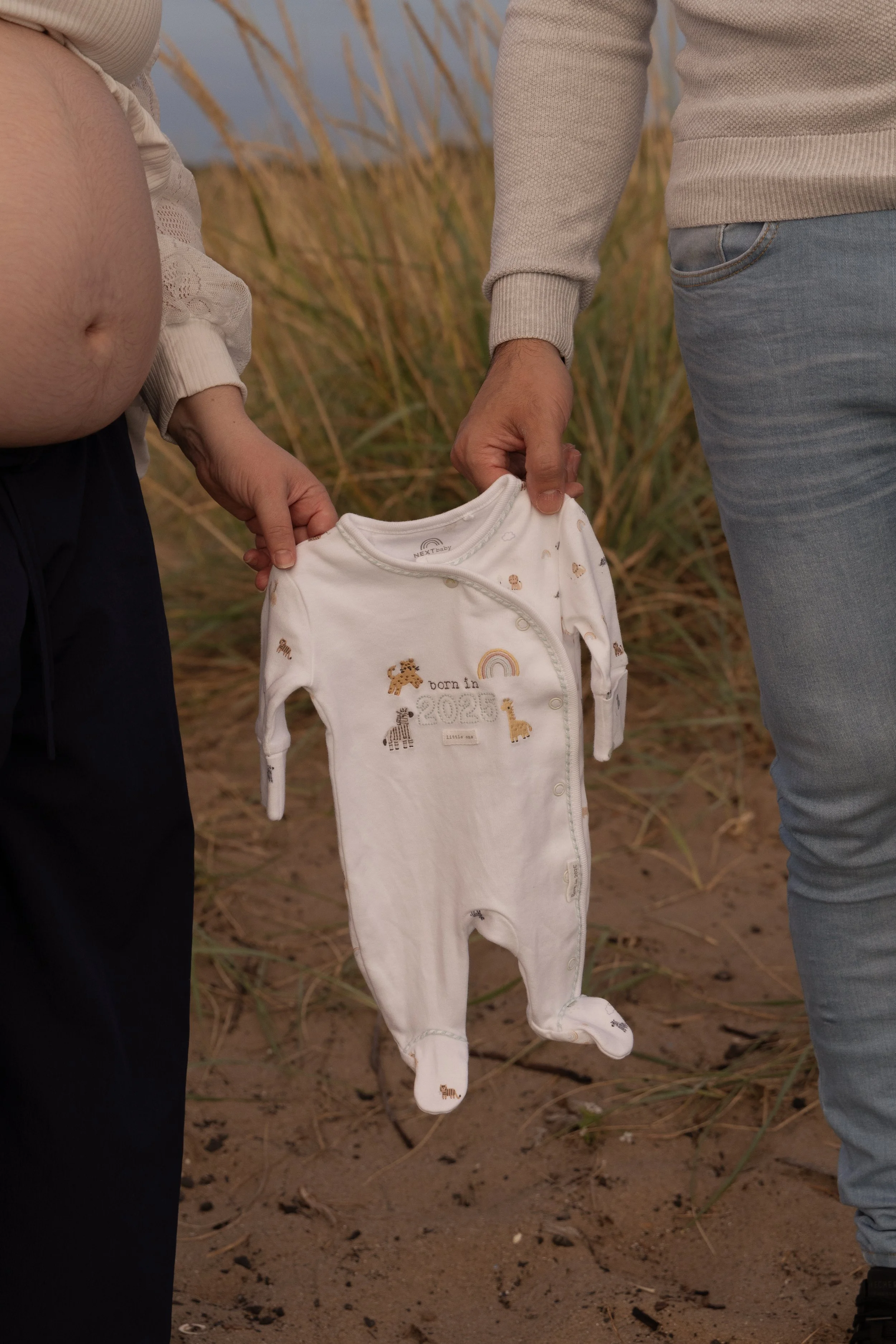 Two people holding a white baby onesie with embroidered animals and the text 'born in 2025', outdoors on sandy ground with tall grass in the background.