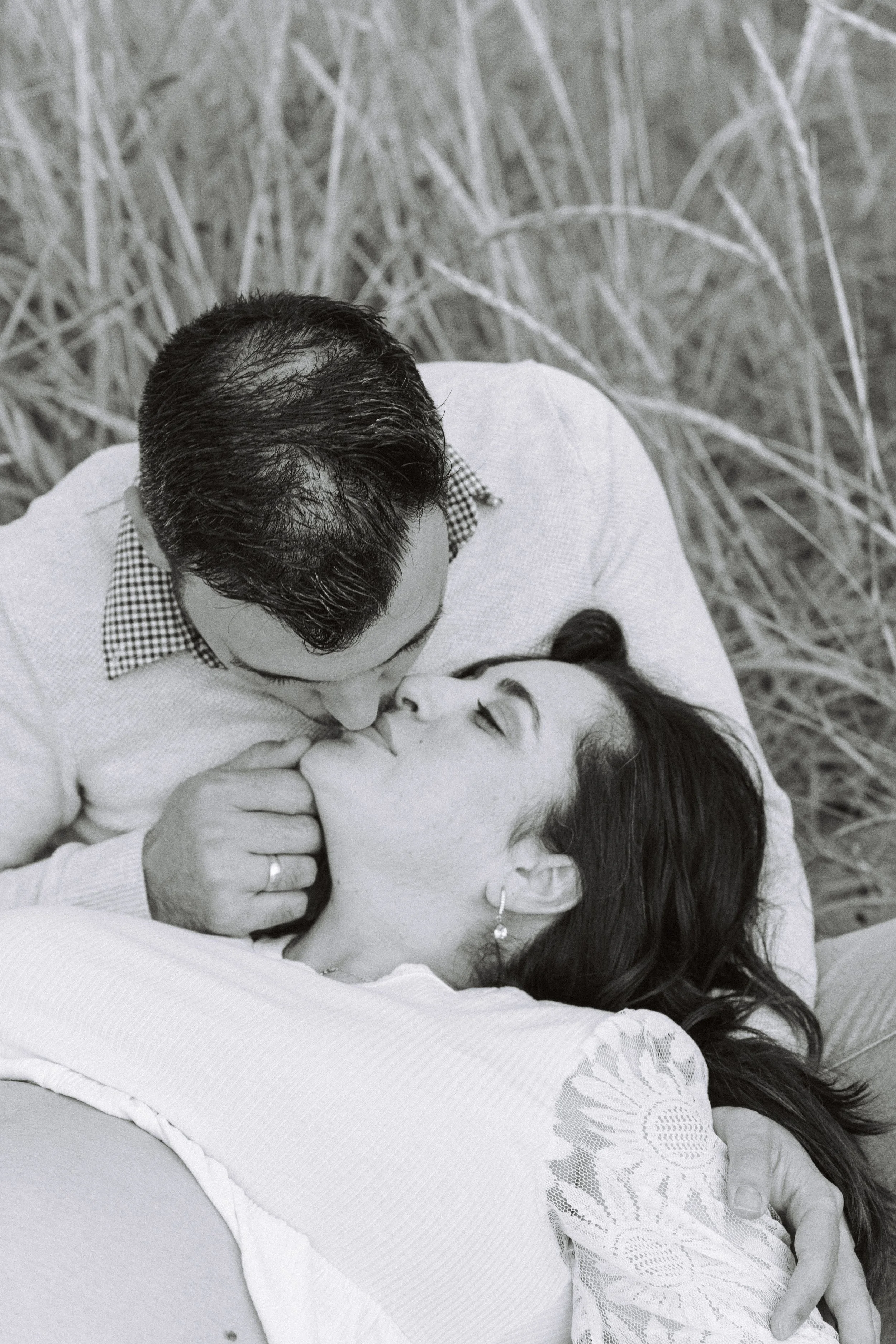A black and white photo of a man and woman in a romantic pose, with the man kissing the woman on the lips while she lies on her back in an outdoor setting with tall grass in the background.
