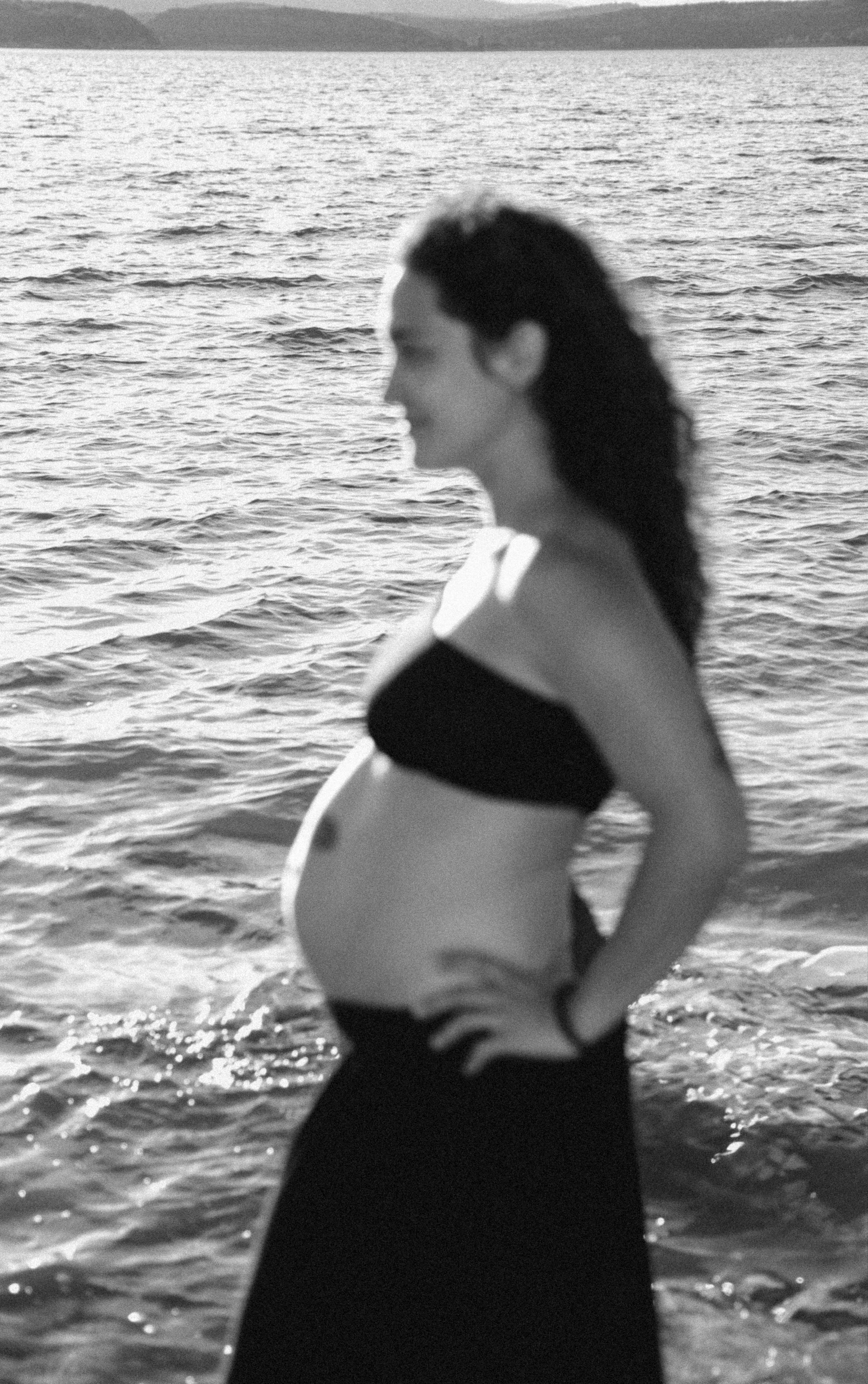 A pregnant woman standing at the edge of the water in a black bikini top and long skirt, facing sideways with her hands on her hips, by a body of water during sunset or sunrise.