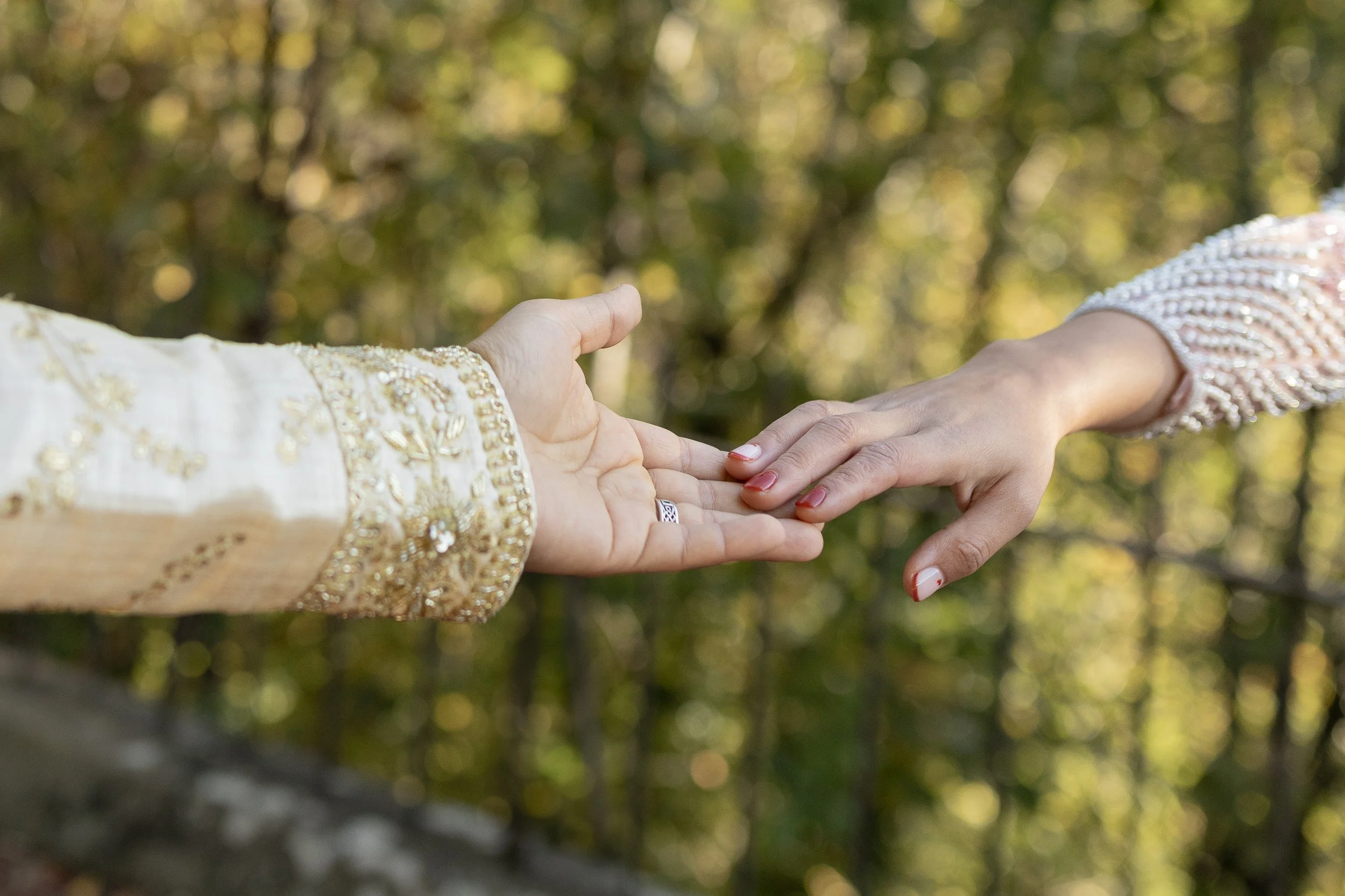 Two people holding hands, with one person reaching out and touching the other's hand. The person on the left wears a cream-colored garment with gold embroidery, and the person on the right wears a pink sleeve with pearl embellishments. The background is blurred with green and yellow foliage.