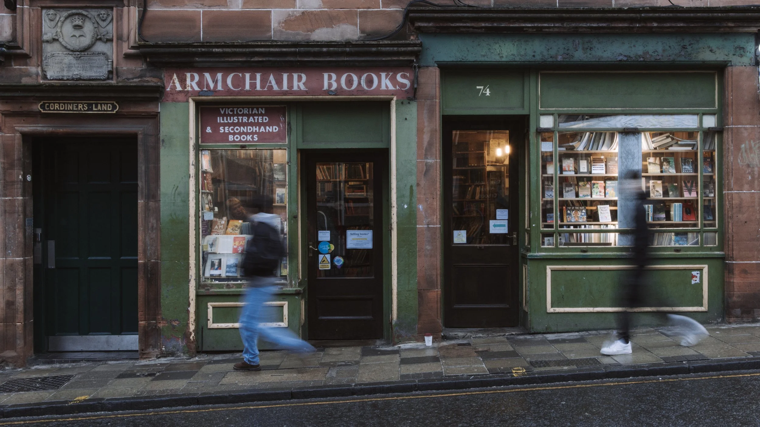 A street view of Armchair Books, a bookstore with green storefront. The window displays many books, and the sign reads 'Victorian Illustrated & Secondhand Books.' The street is wet, and two blurred pedestrians walk past.