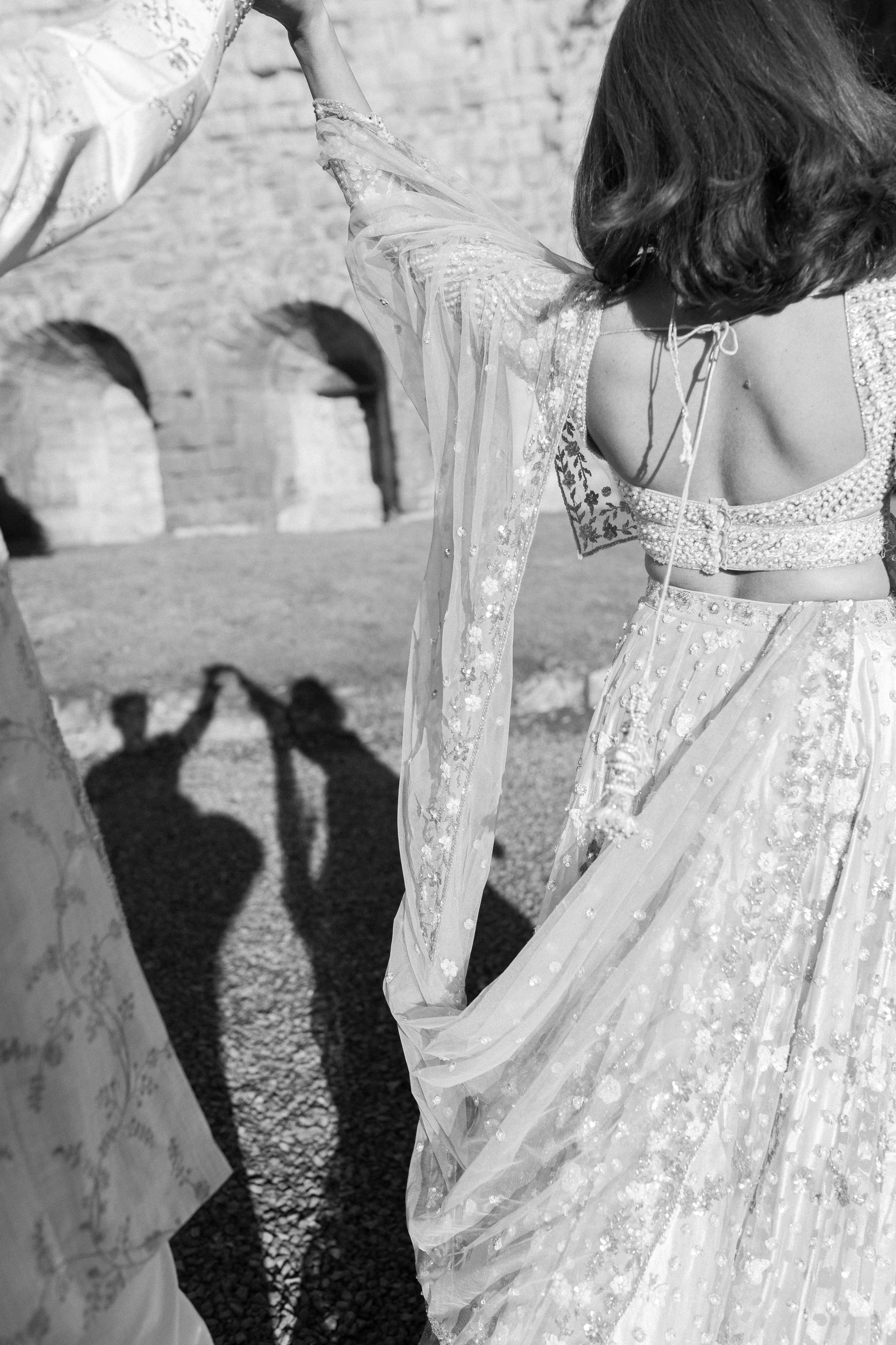 A woman in a traditional Indian outfit, possibly a saree or lehenga, with intricate embroidery, standing outdoors near ancient stone arches. Her shadow is cast on the ground, showing her holding on to someone's hand.