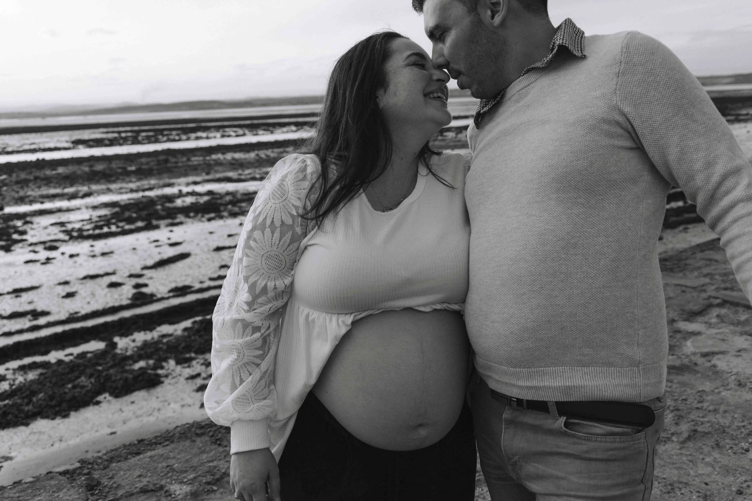 A pregnant woman and a man on a beach in Scotland sharing a tender moment, their foreheads touching, in black and white.