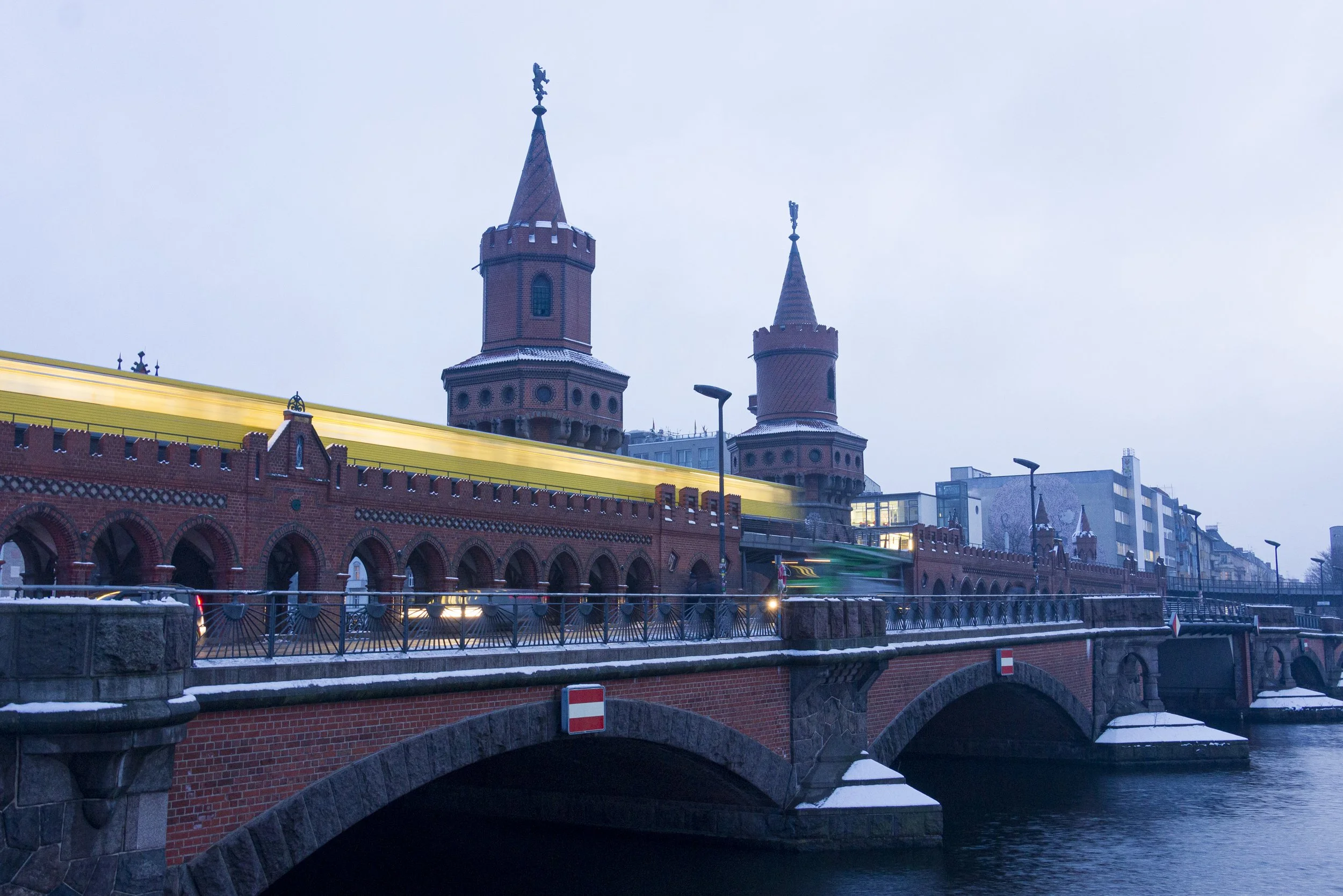 A historic red brick bridge with two towers over a river, modern city buildings in the background, and light snow on the bridge and surrounding areas, during dusk.