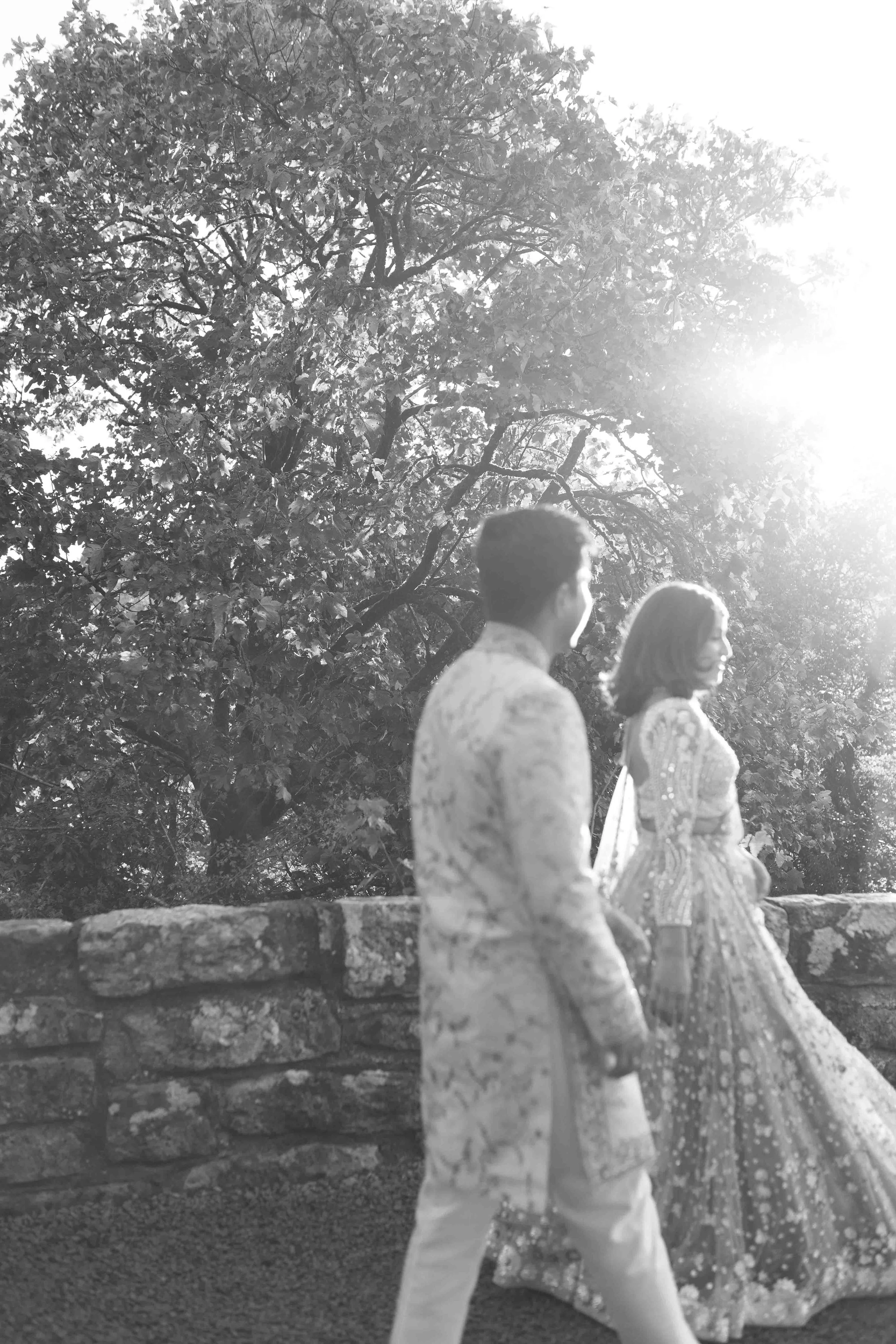 A black and white photo of a man and woman in traditional wedding attire standing outdoors on a stone wall with trees and sunlight in the background.