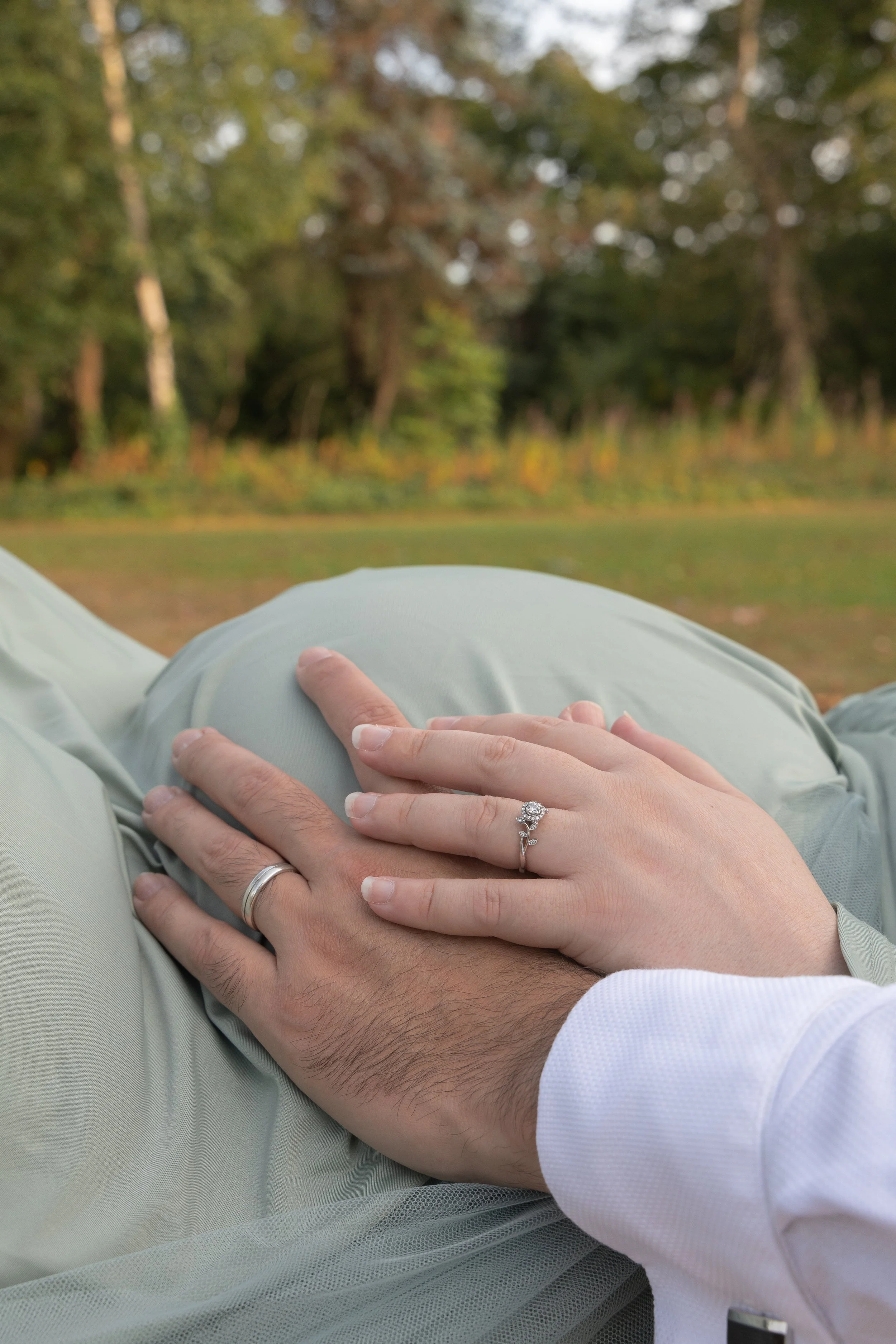 Close-up of a couple's hands with wedding rings resting on a person's pregnant belly in an outdoor setting with trees and greenery in the background.