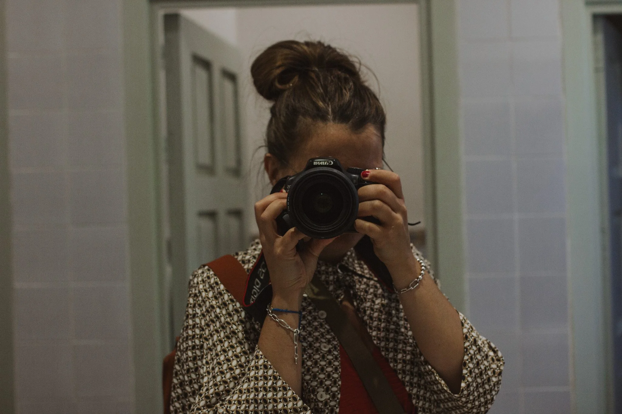 Person taking a selfie with a camera in front of a mirror, with a bun hairstyle, in a bathroom with white tiled walls.