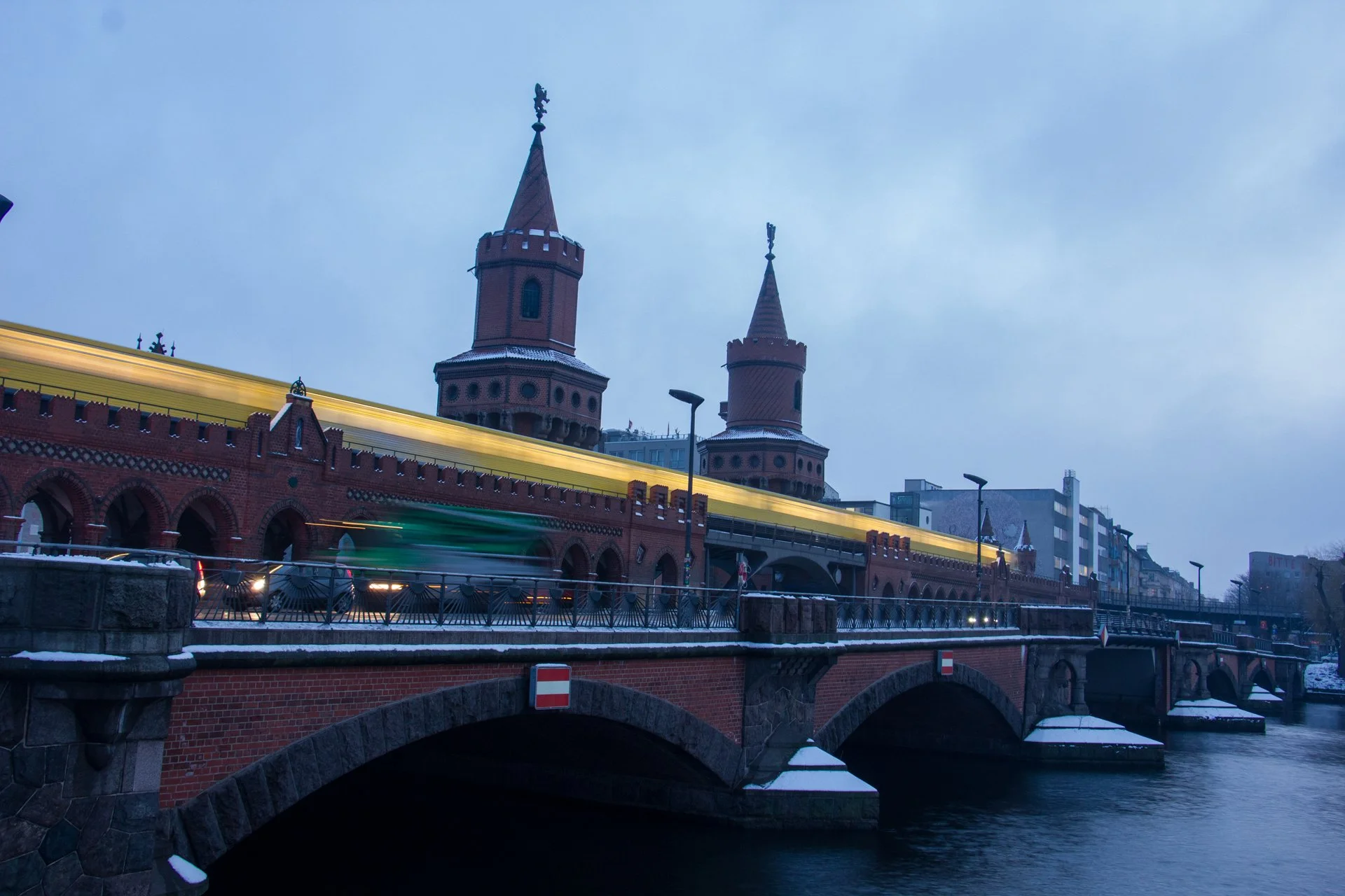 A historical brick bridge over a river with two castle-like towers in the background and a train passing on the bridge at dusk.