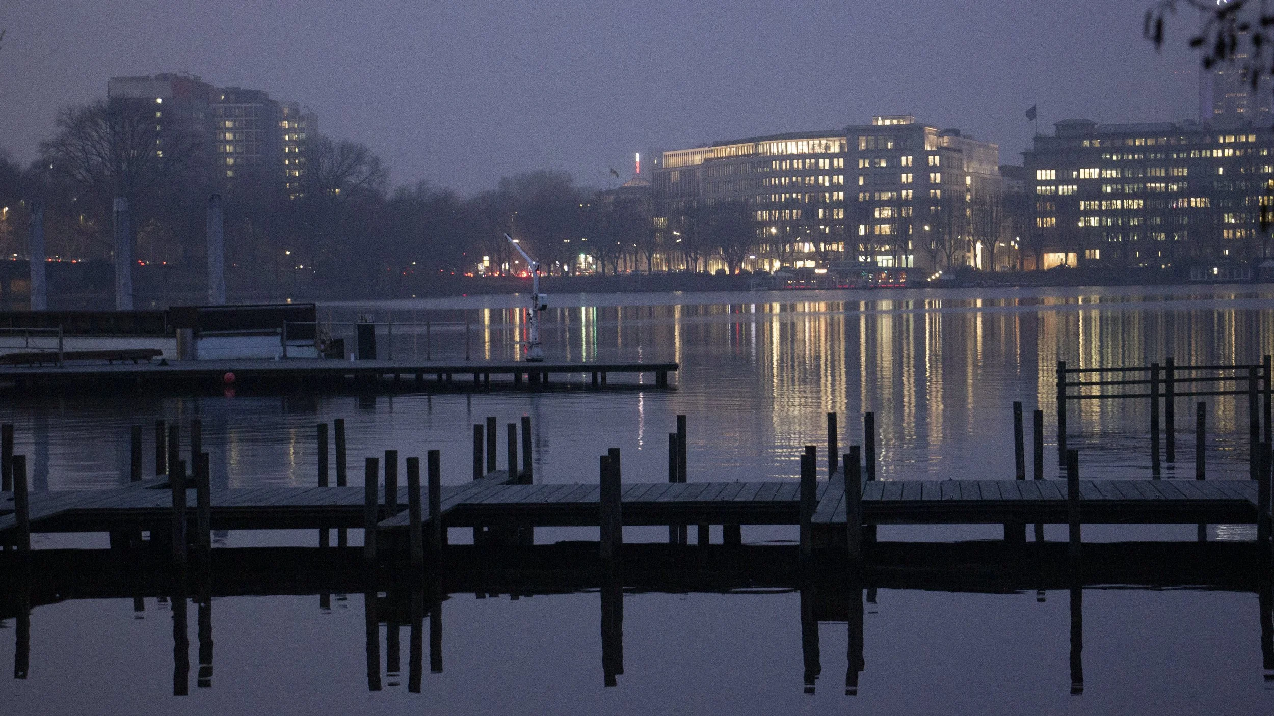 Nighttime view of a calm river with a dock in the foreground, modern buildings with lights on across the river, and trees along the shoreline.