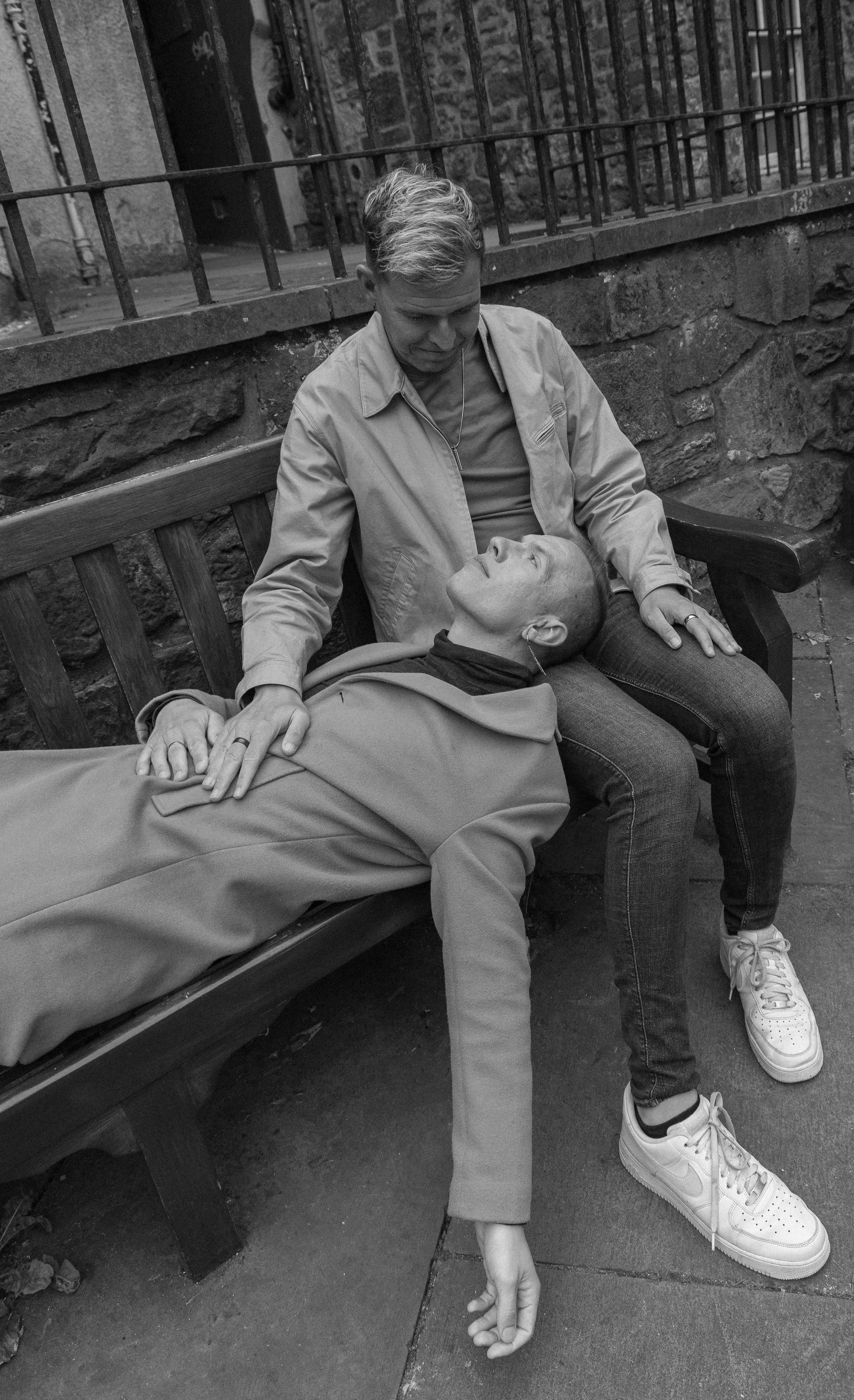 Two women sit on a park bench; one is lying down with eyes closed, and the other is sitting and gently holding her hand and shoulder, showing care and concern.