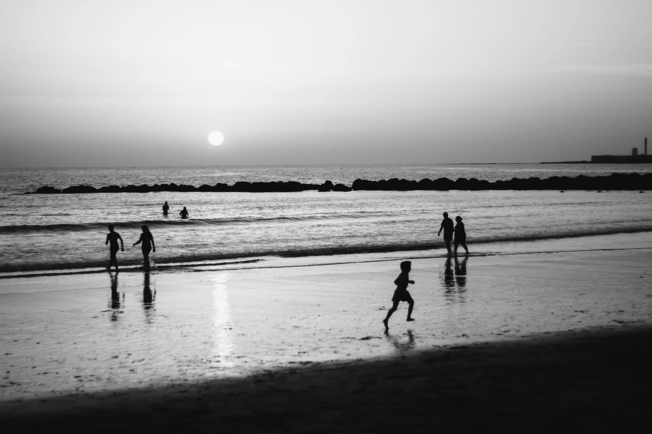 Silhouettes of children and adults on a beach at sunset, with calm waves, rocky breakwater, and distant buildings in the background