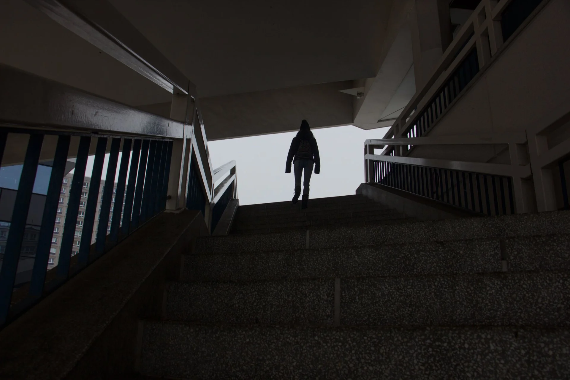 A silhouette of a person walking up stairs in a multi-story building, viewed from below with the open sky in the background.