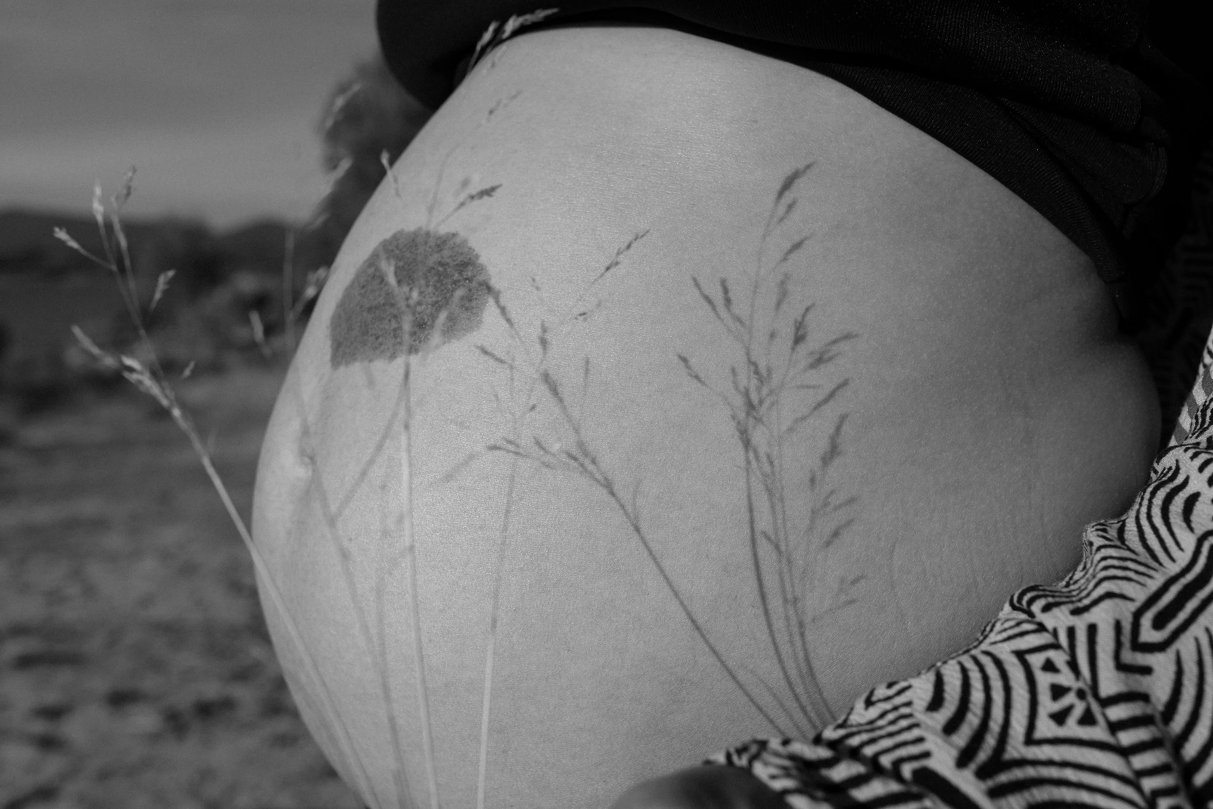 Fine art Close-up of pregnant woman with a tattoo of wildflowers on the thigh, in a black and white outdoor setting.