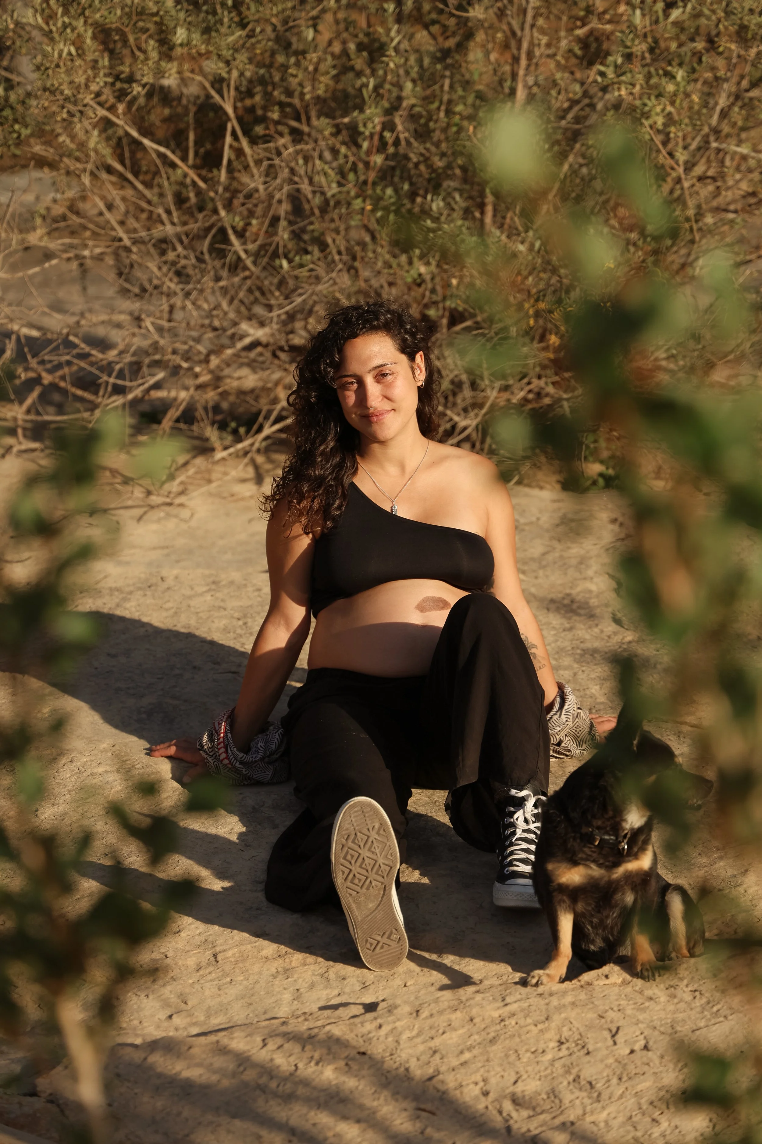 A woman with curly dark hair in a black one-shoulder crop top and black pants sitting on sandy ground outdoors with a dog beside her, vegetation behind and in front, during sunlight.