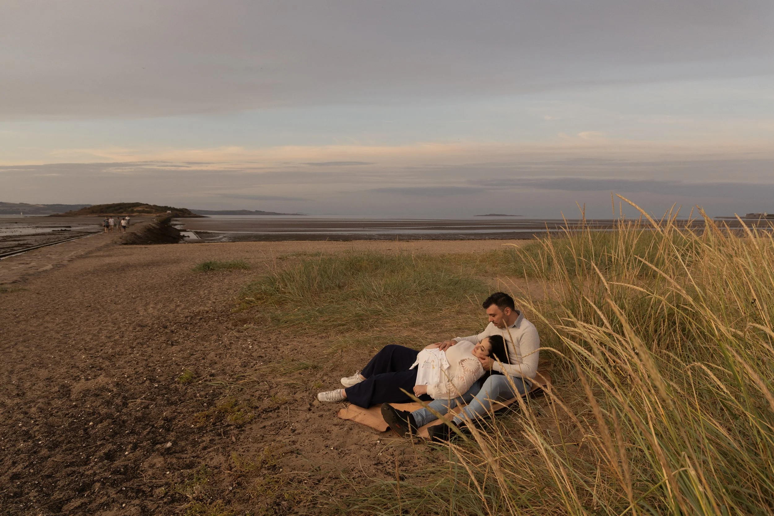 A couple sitting and lying in the grass on a beach at sunset, with a few people walking on the sand in the background.