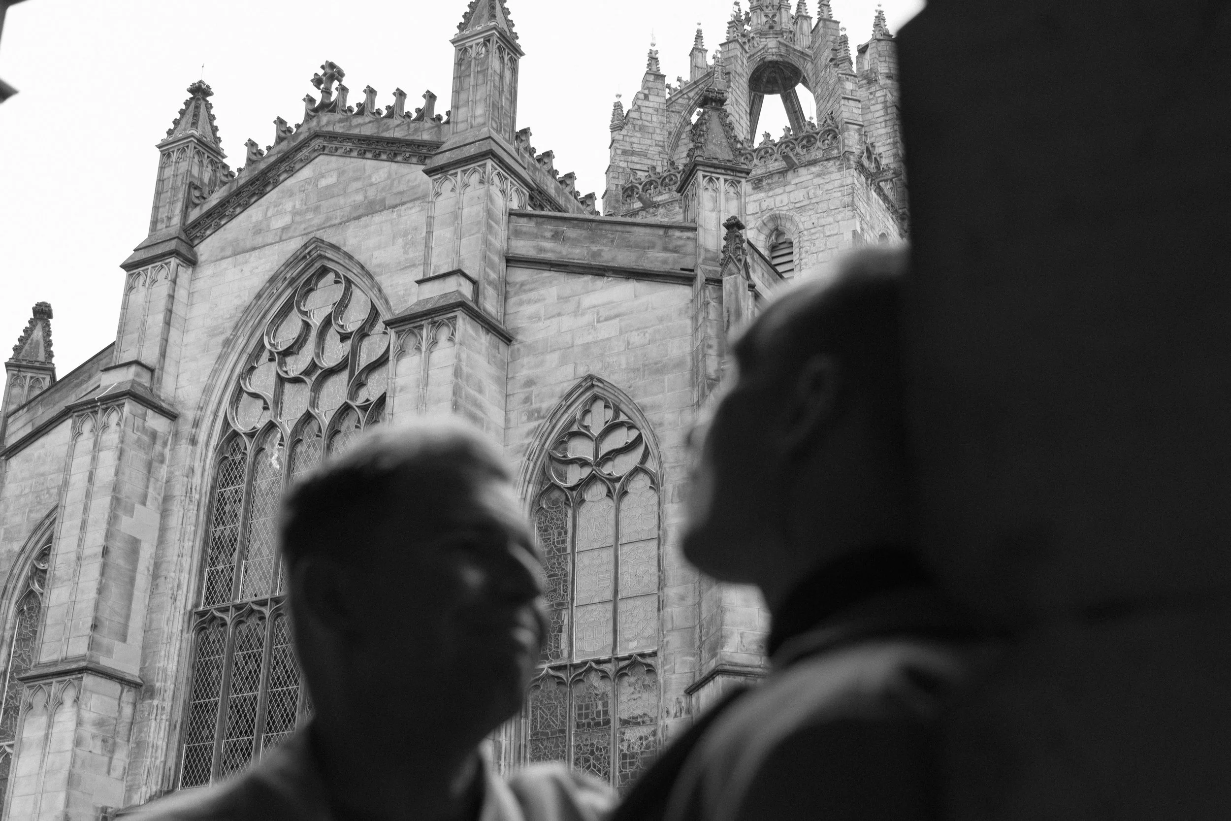Silhouettes of two people, blurred in the foreground, with a Gothic-style cathedral with ornate windows and spires in the background, whihc is St Giles cathedral in Edinburgh