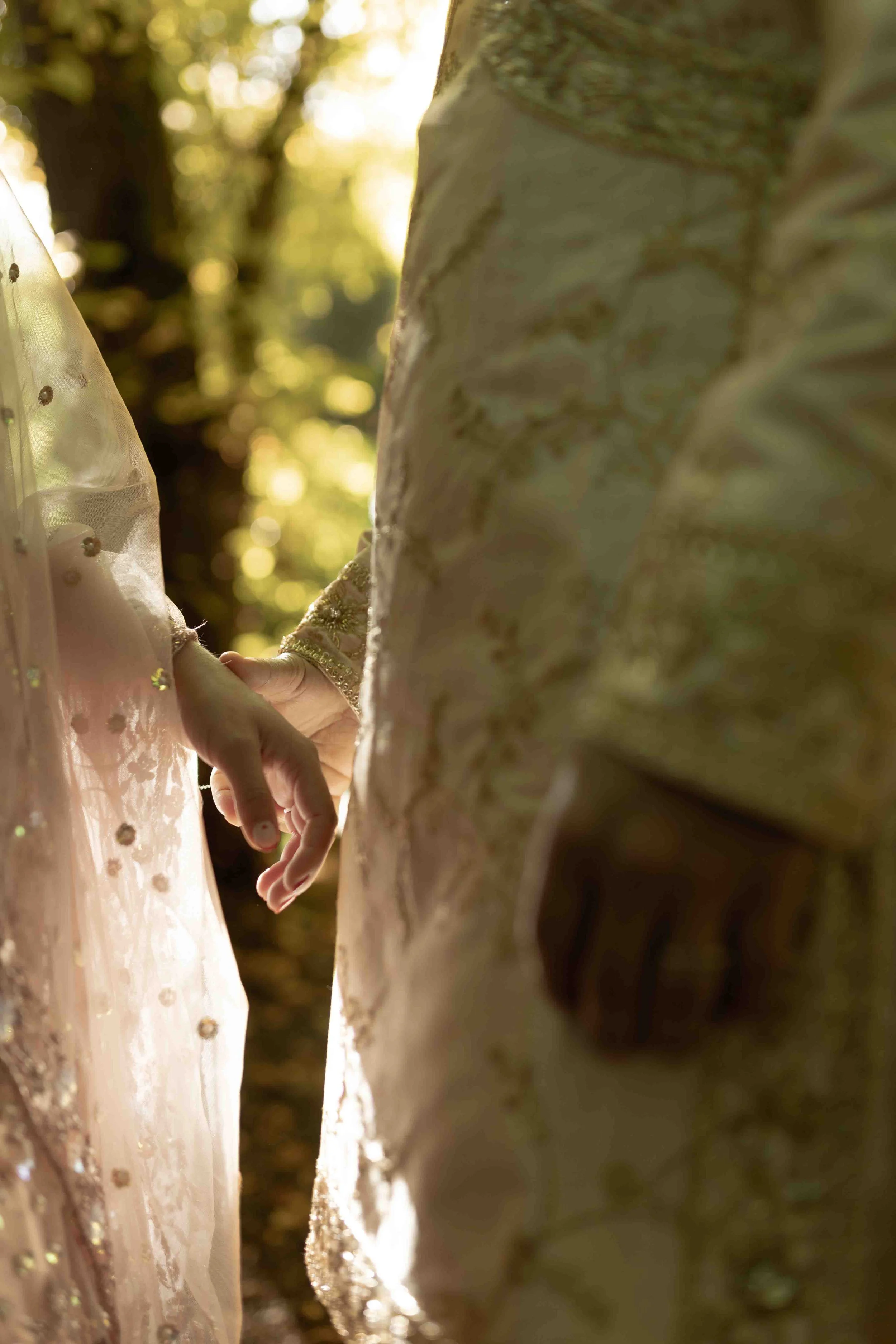 Close-up of two people holding hands, partially visible, in a sunlit outdoor setting with trees in the background.