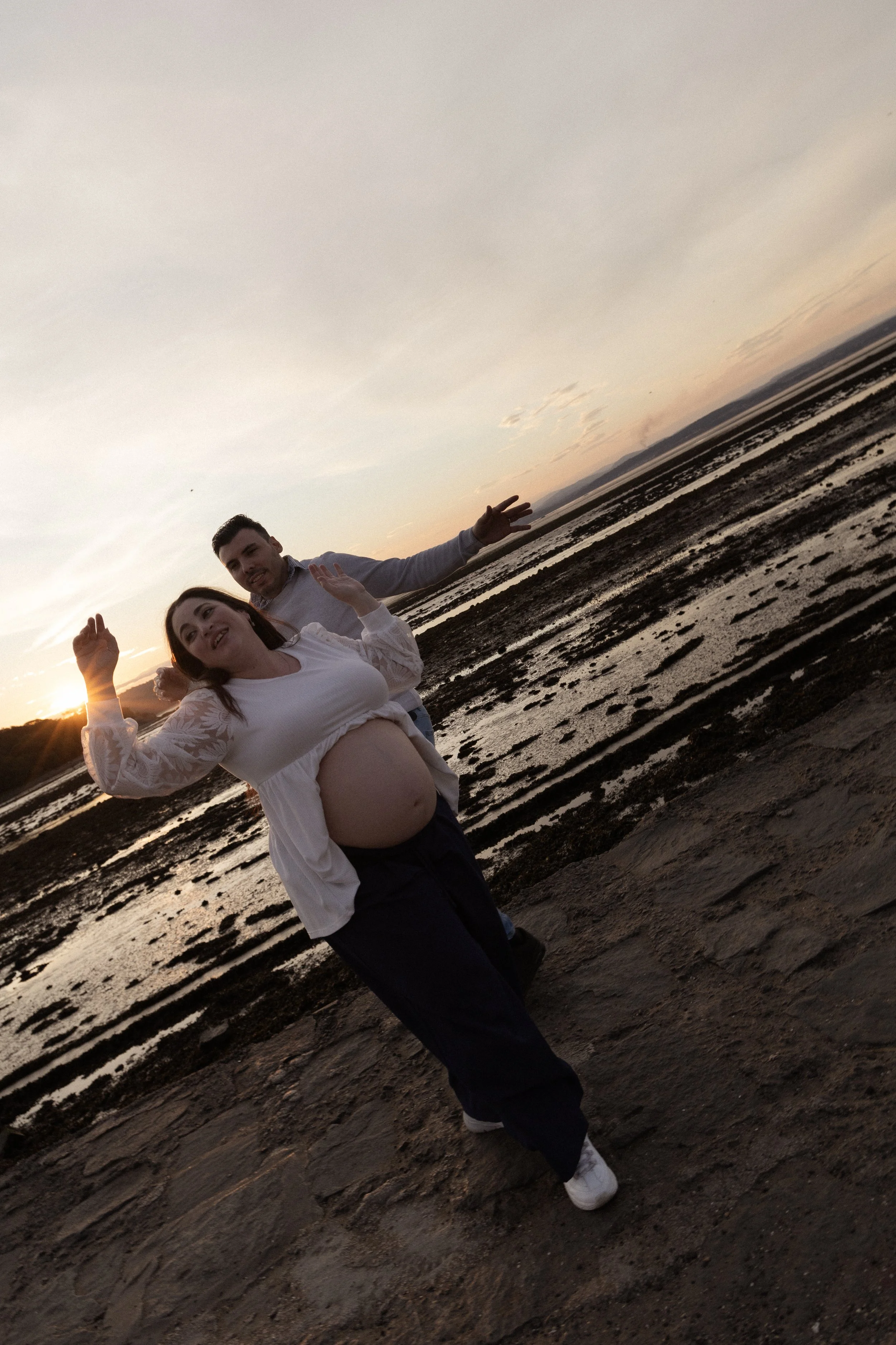 A pregnant woman and a man at the beach during sunset, with the woman smiling and the man smiling, standing on rocks with the sea in the background.