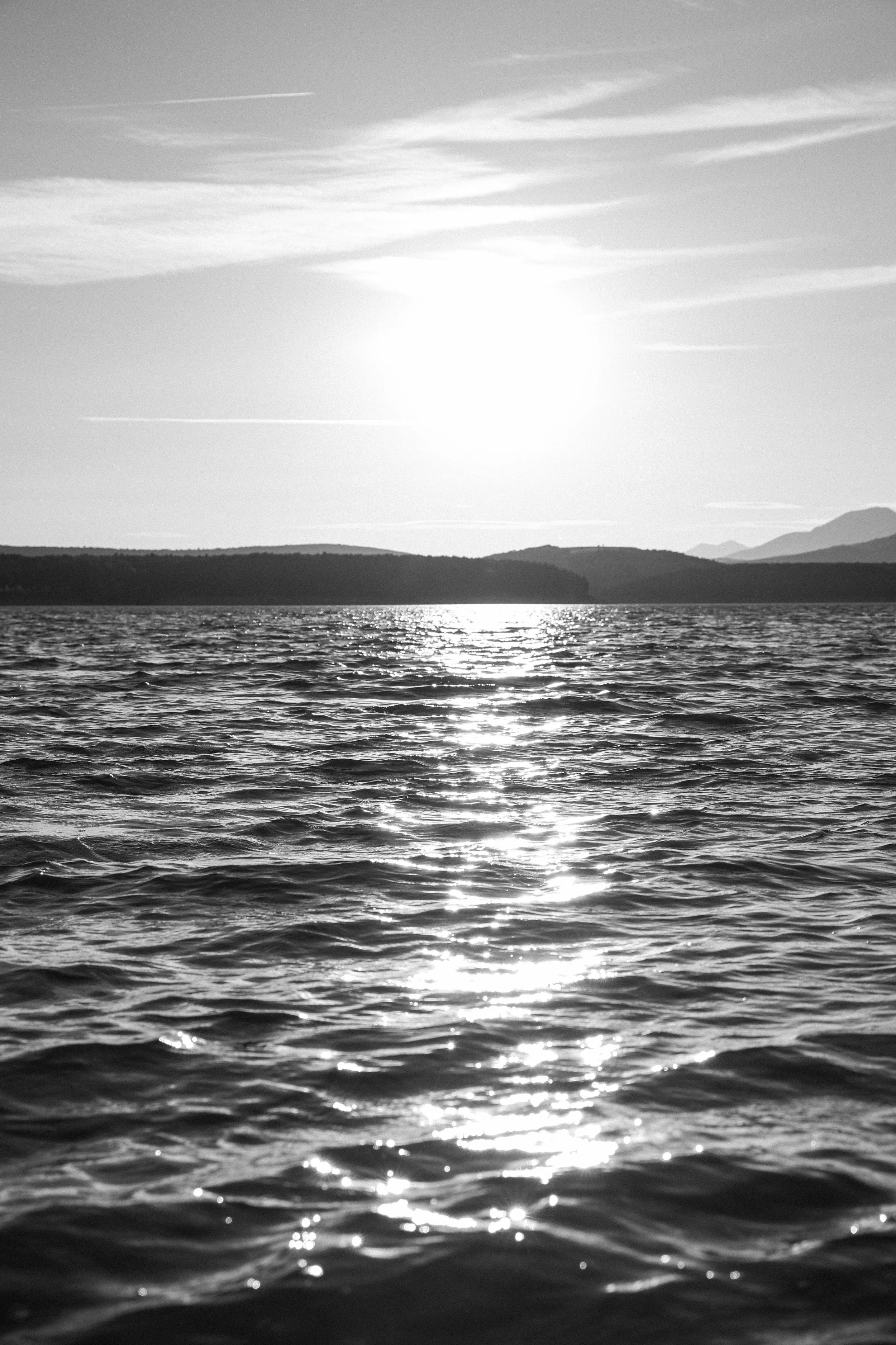 Black and white photo of a body of water with ripples, reflecting sunlight, with land and mountains in the background under a sky with wispy clouds.