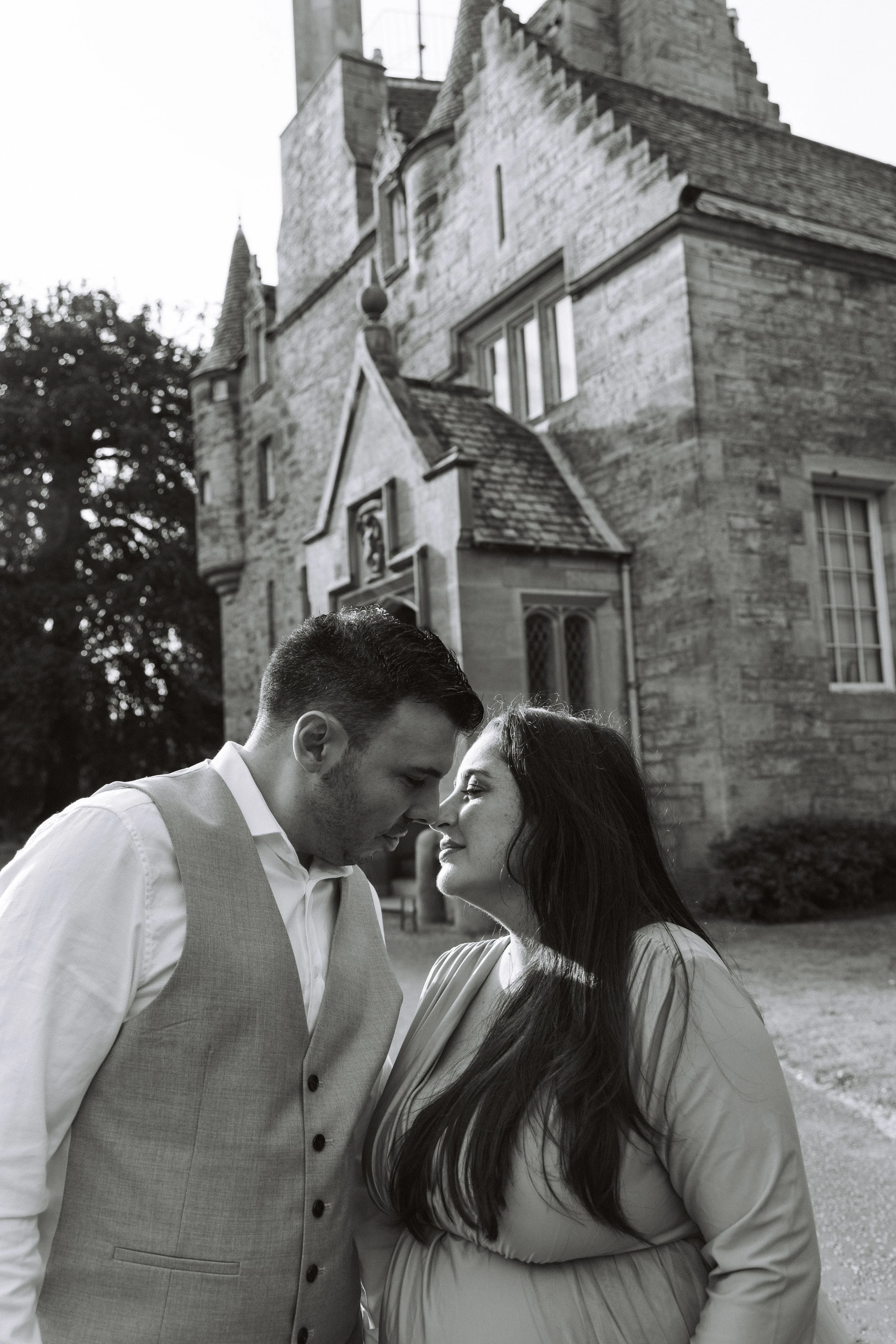 A black and white photo of a couple with foreheads touching, standing close outside a historic stone building. The man is wearing a white shirt and vest, and the woman is pregnant, wearing a long-sleeve top.