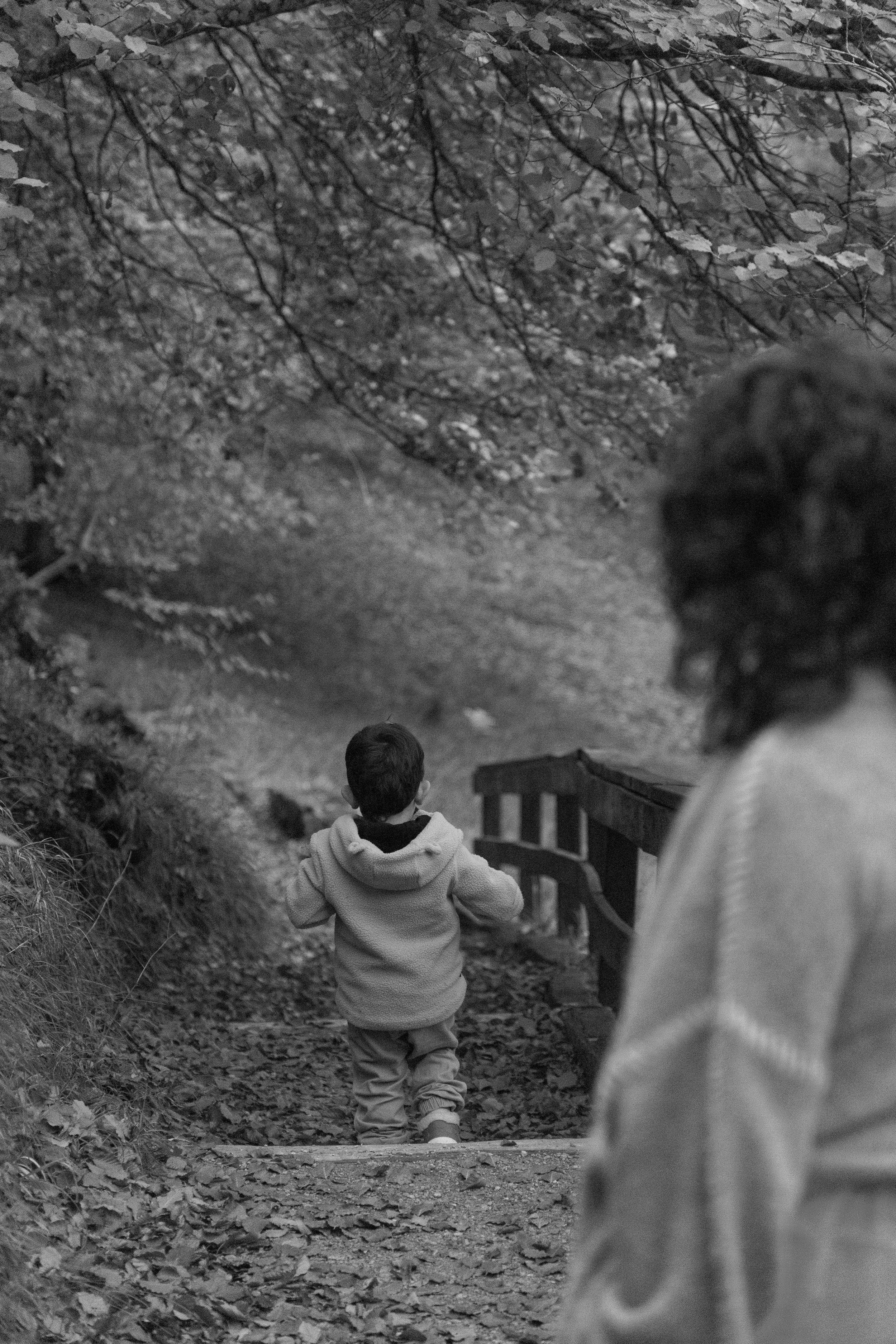 A boy and a woman are walking down a forest trail surrounded by trees and fallen leaves.
