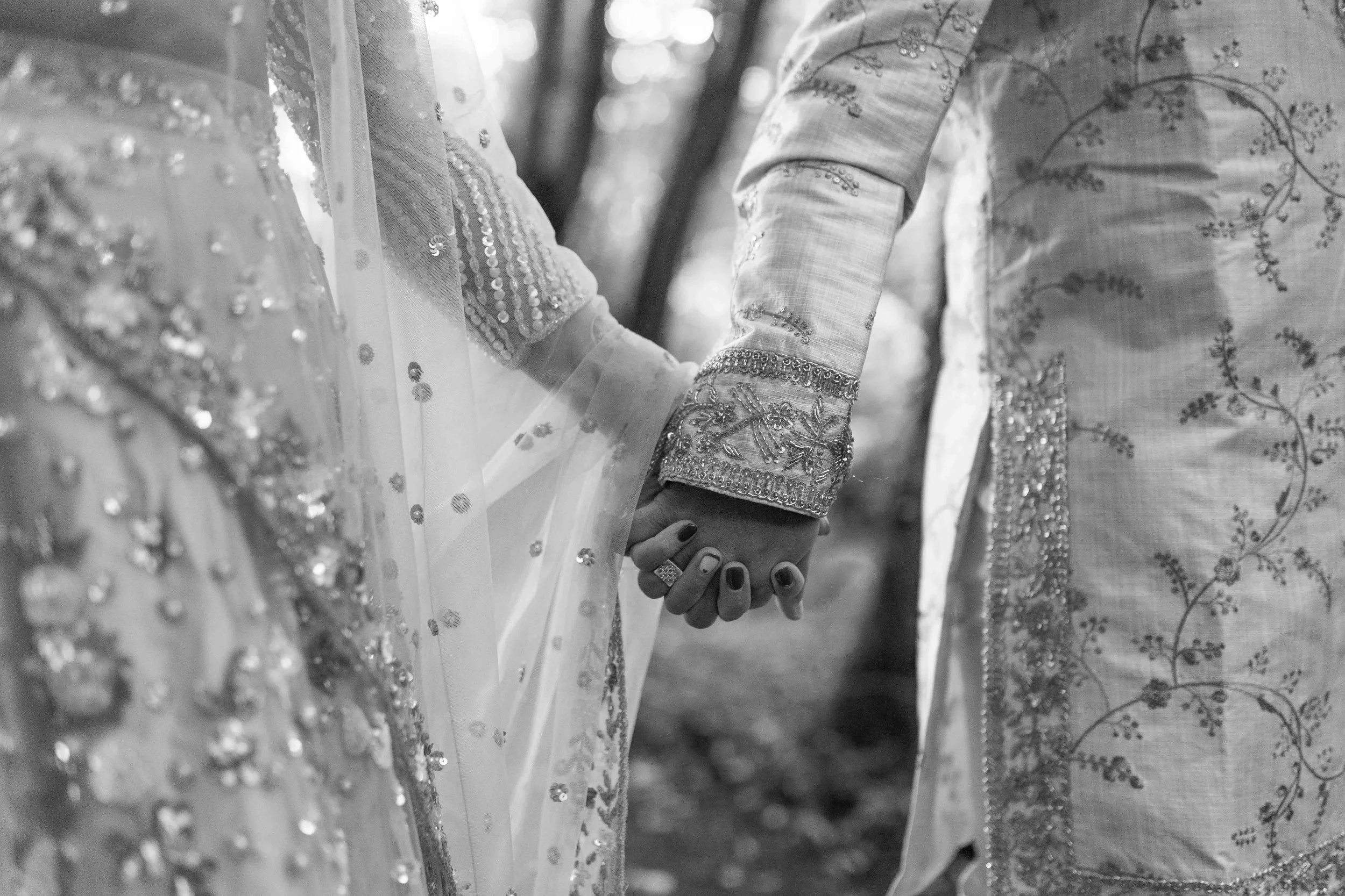 A close-up of a couple holding hands, dressed in traditional wedding attire, with the focus on their joined hands in a natural outdoor setting.