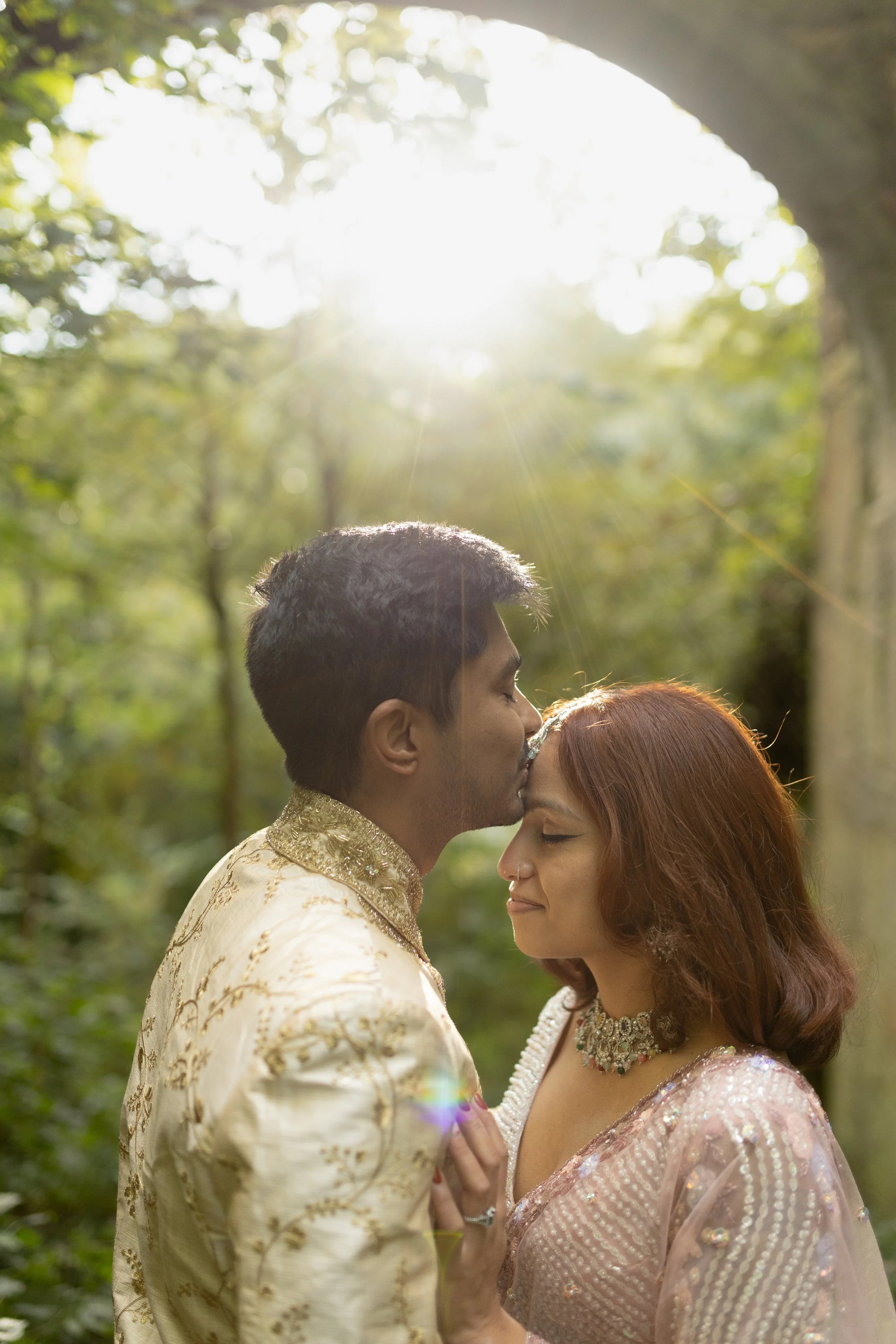 Couple dressed in traditional attire sharing an intimate moment in a sunlit forest.