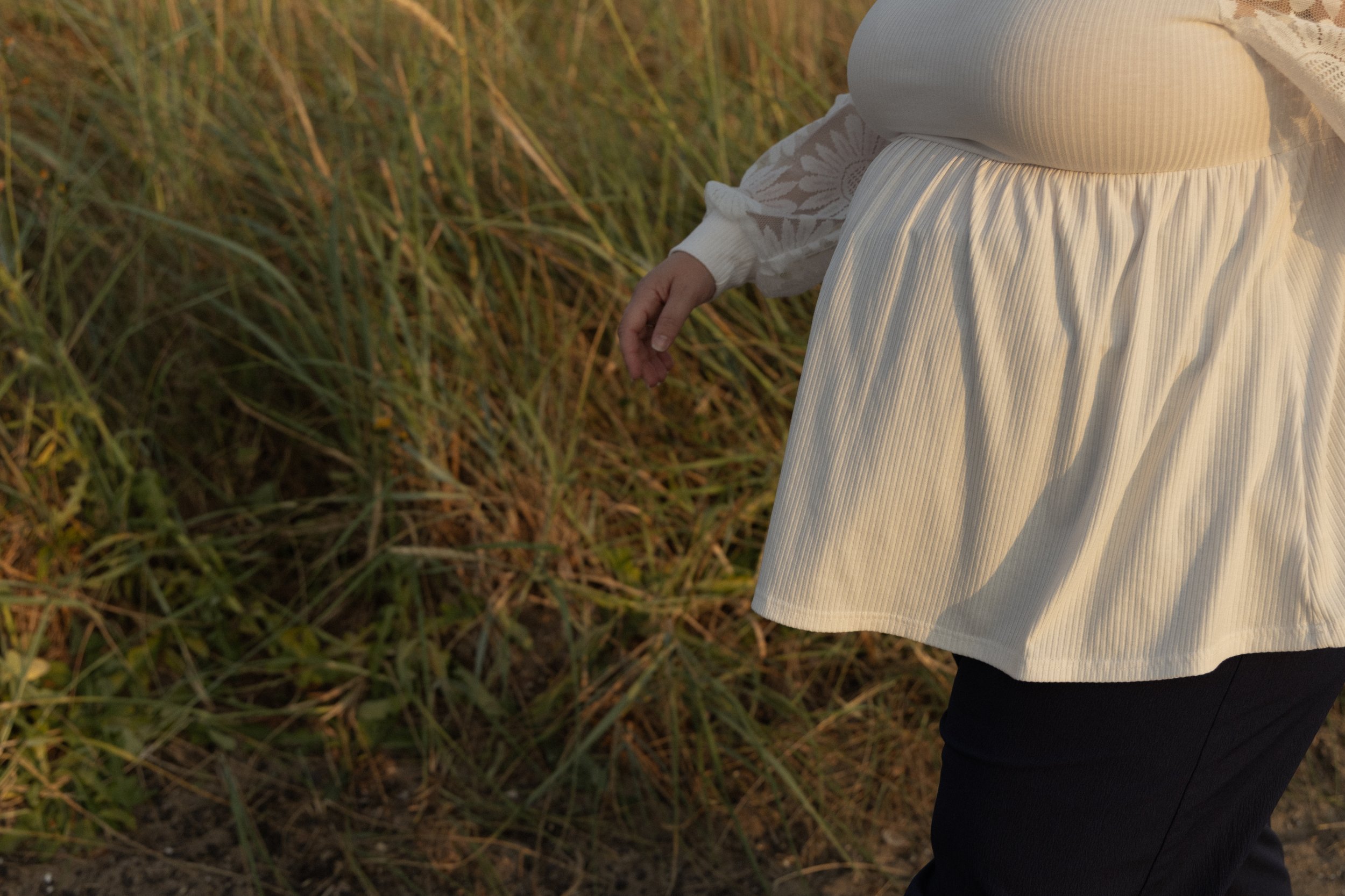 Close-up of a person wearing a white textured dress and black pants, walking through a grassy field during sunset.